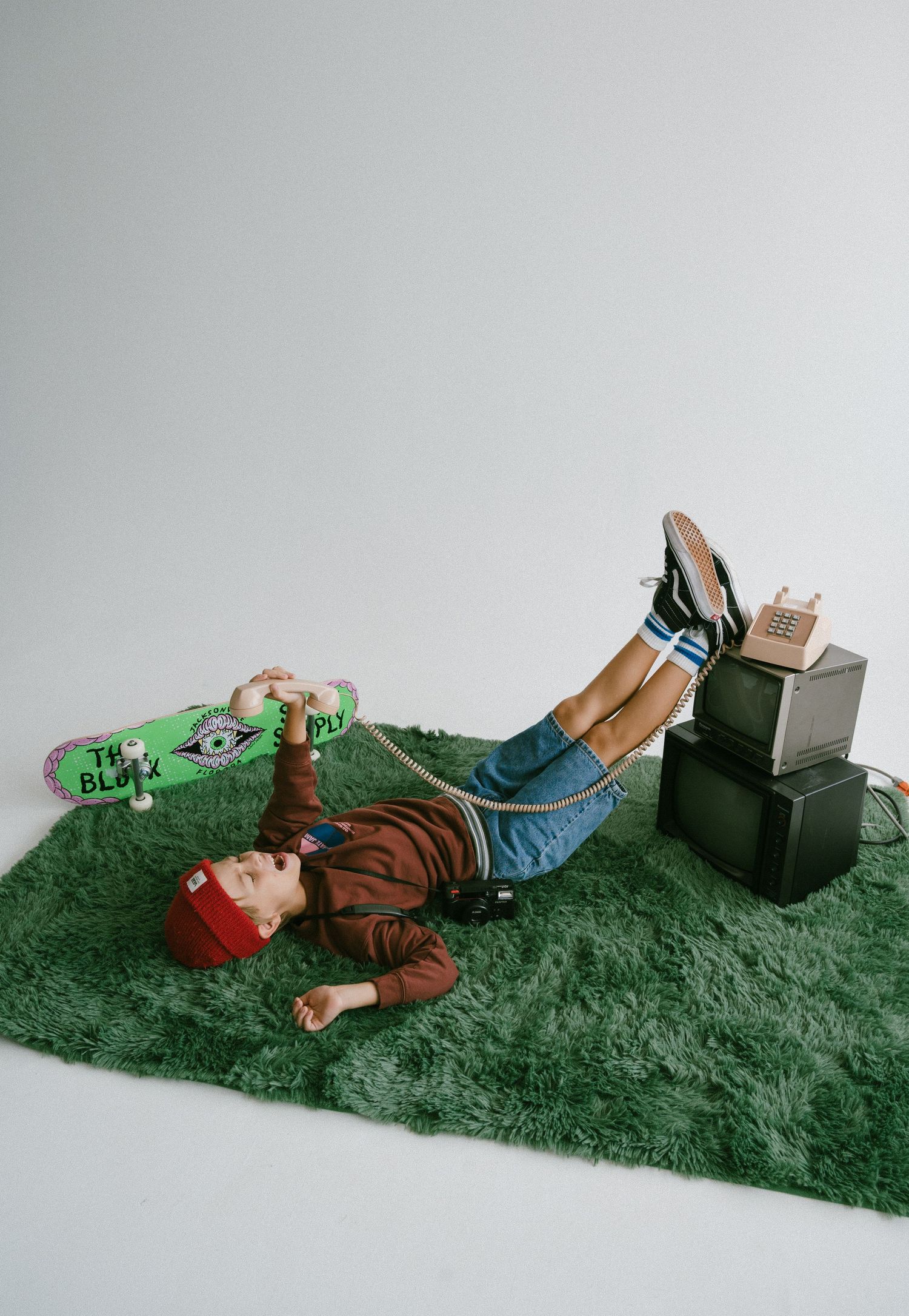 Young man lying on green carpet rug with a skateboard and vintage electronics in a studio setting.
