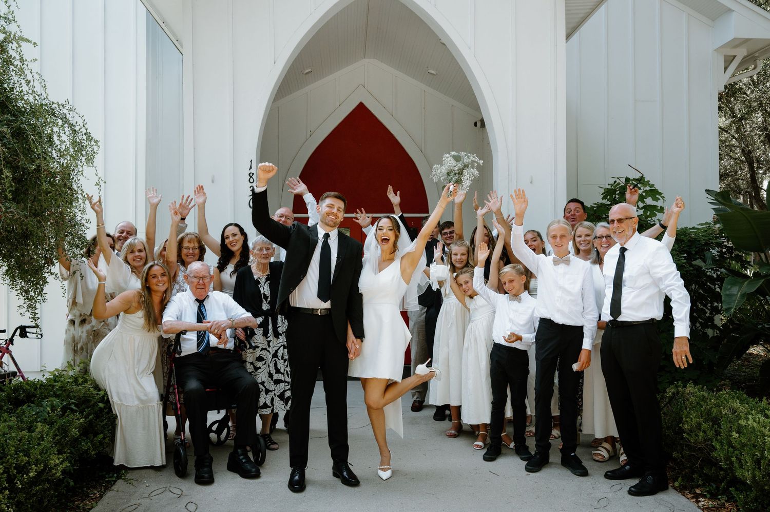 Wedding party celebrating outside white chapel with red doors, hands raised in joy after ceremony on Amelia Island, Florida.