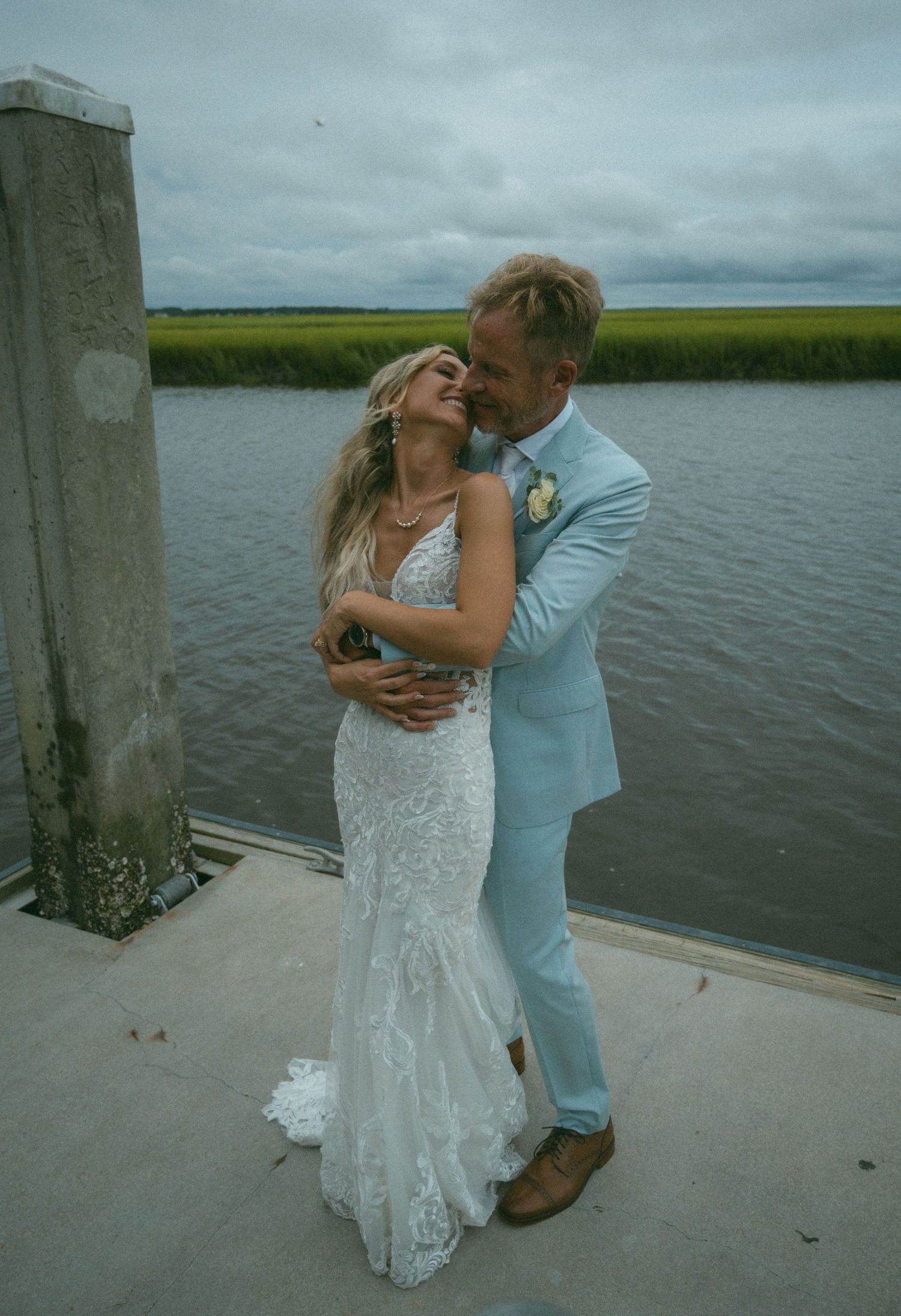 Bride and groom share a romantic embrace on a waterfront dock, with marsh views behind them.