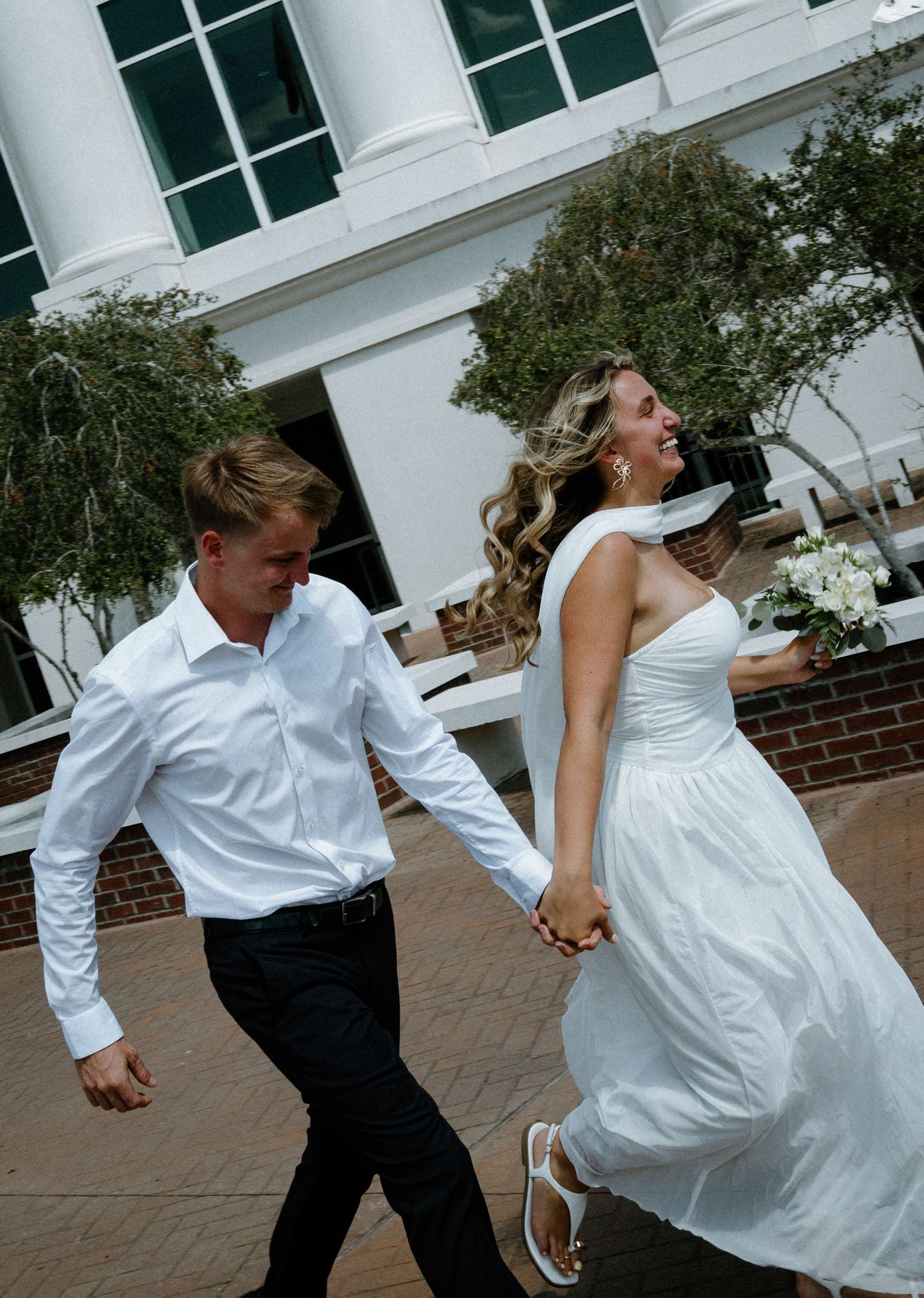 Joyful bride in white dress holding bouquet runs hand-in-hand with groom in white shirt outside brick building in Fernandina Beach, Florida.