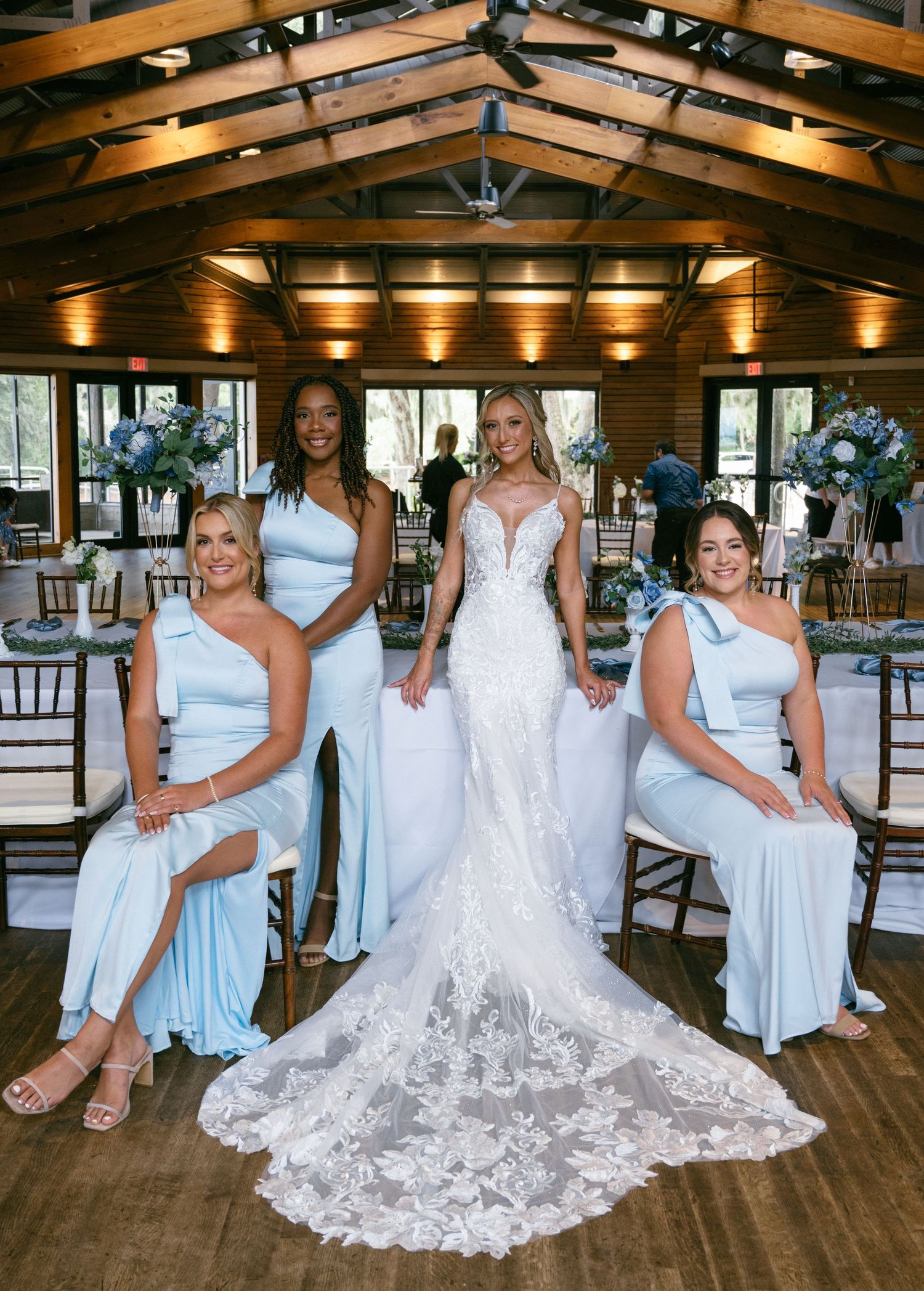 Bride in lace gown poses with three bridesmaids in light blue dresses inside a rustic wooden venue.