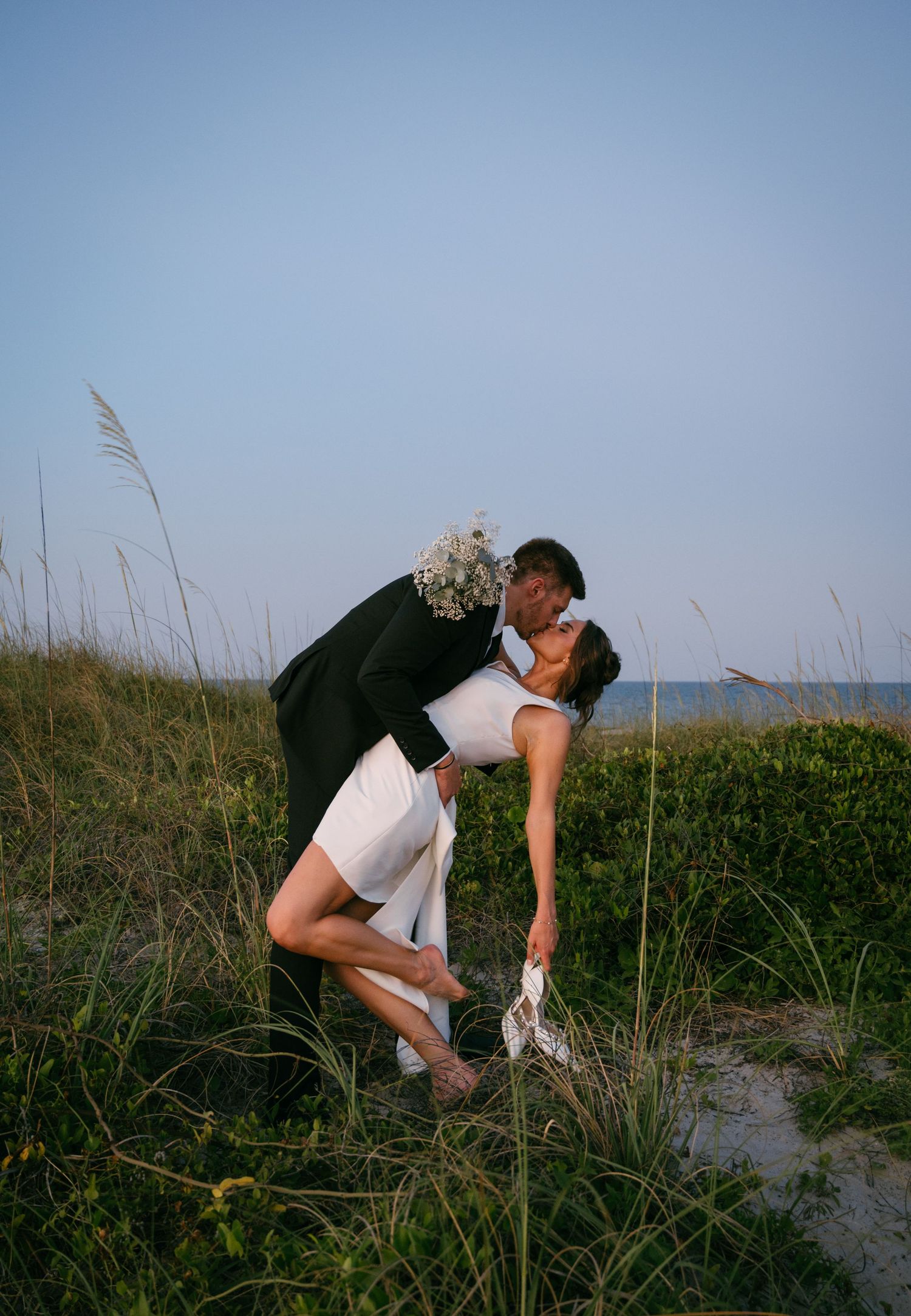 Groom dips bride for romantic kiss among sea oats on sandy beach at sunset at the Ocean Club of Amelia.