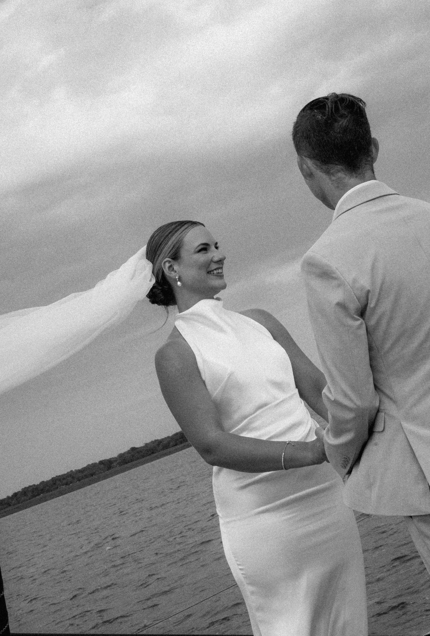 Black and white photo of smiling bride in white gown with flowing veil facing groom outdoors.