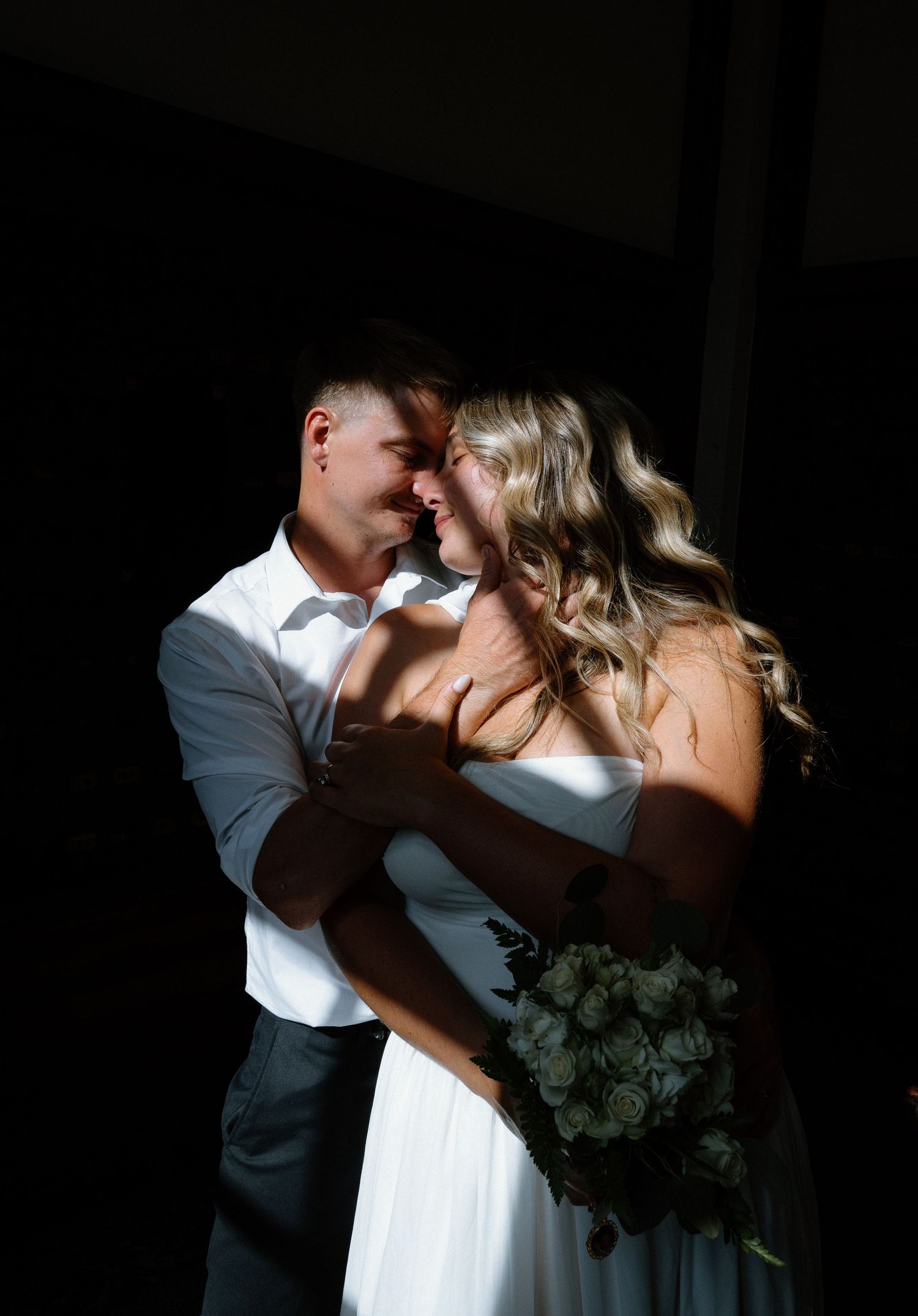 Romantic newlywed couple embracing in dramatic sunlight and shadow with bride holding white bouquet.