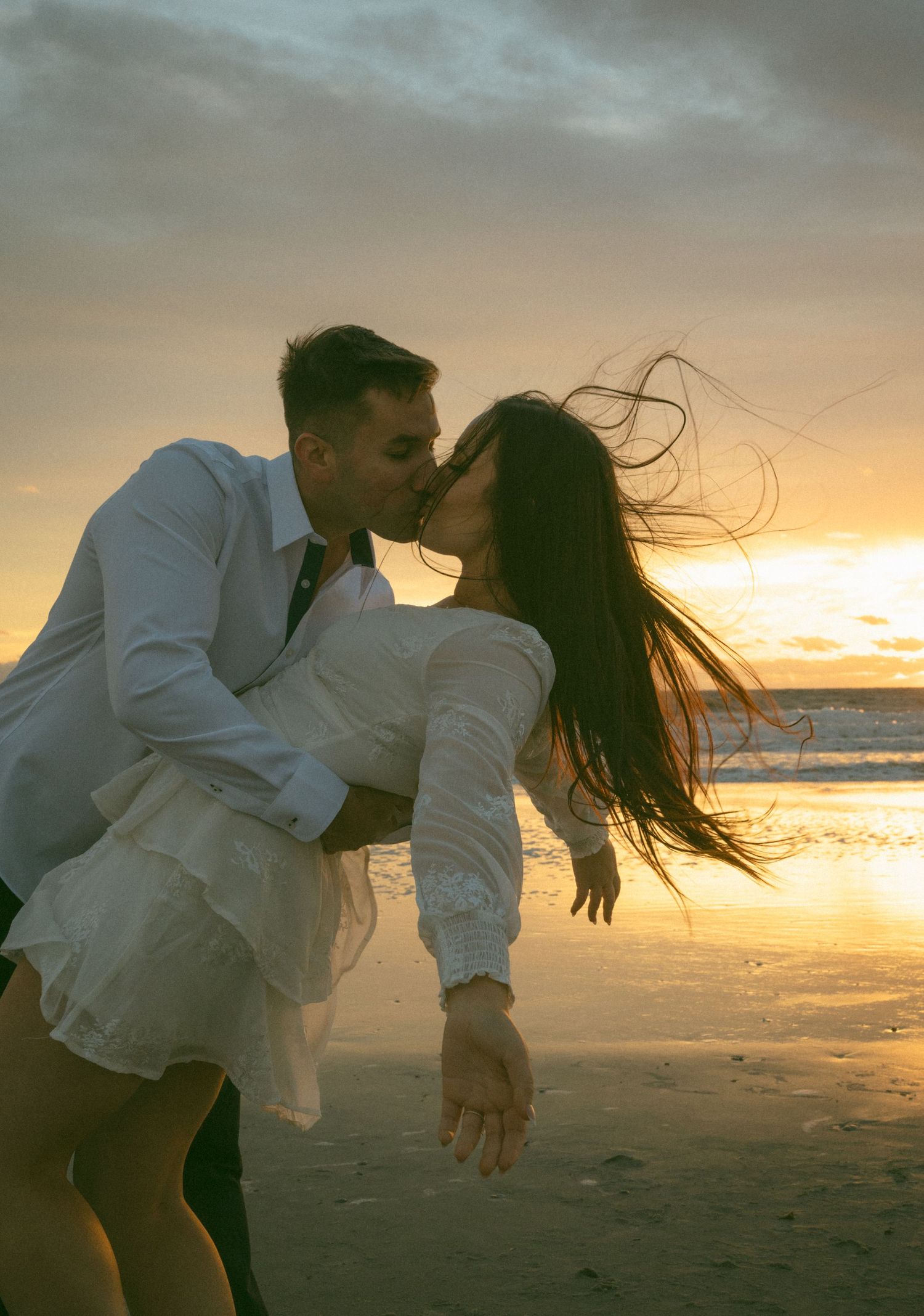 Couple sharing a romantic dip kiss on the beach at golden sunset with wind-blown hair on Amelia Island, Florida.