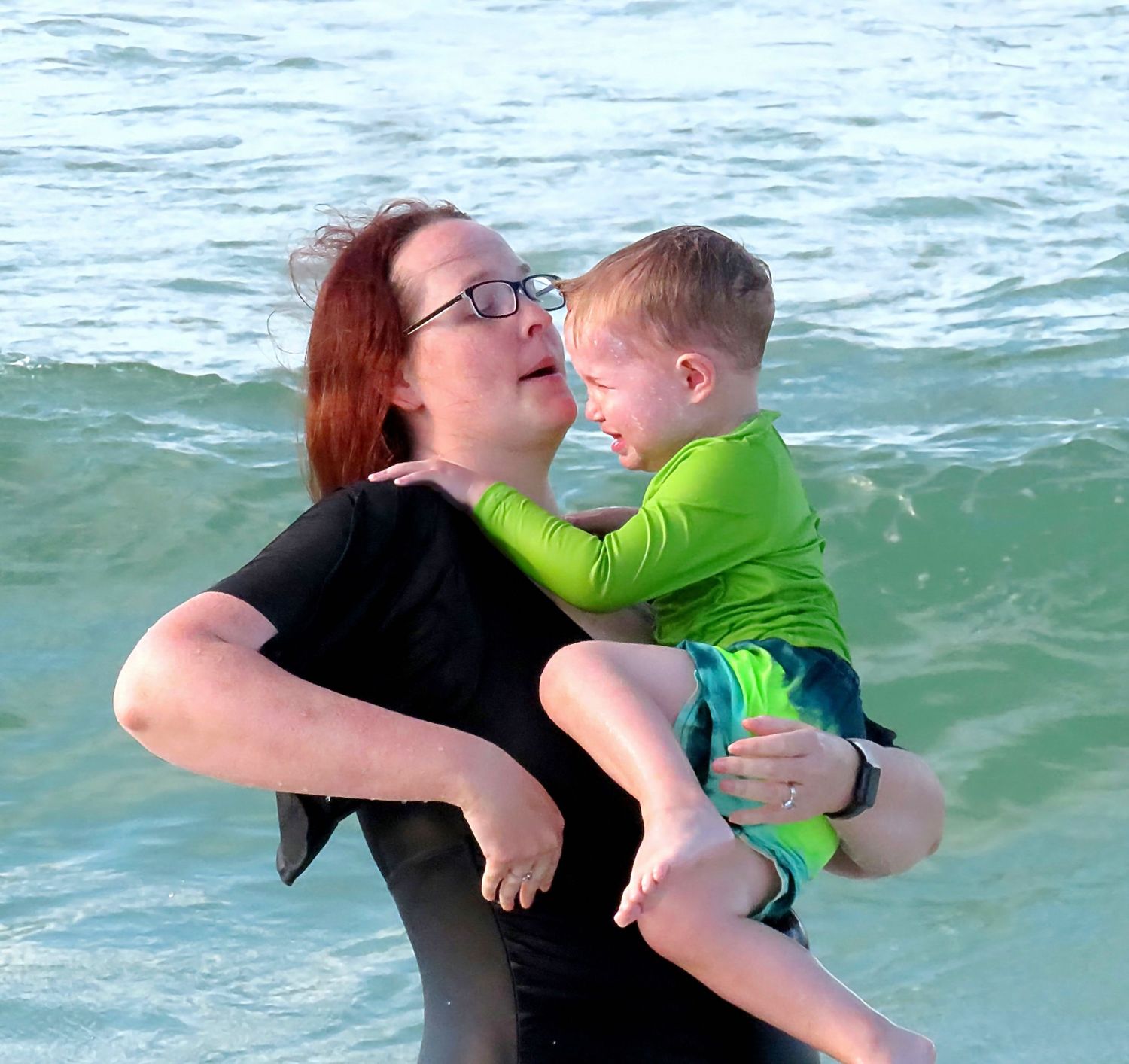 Red-haired woman in black swimsuit holds a crying toddler boy in green rashguard at the ocean shore.