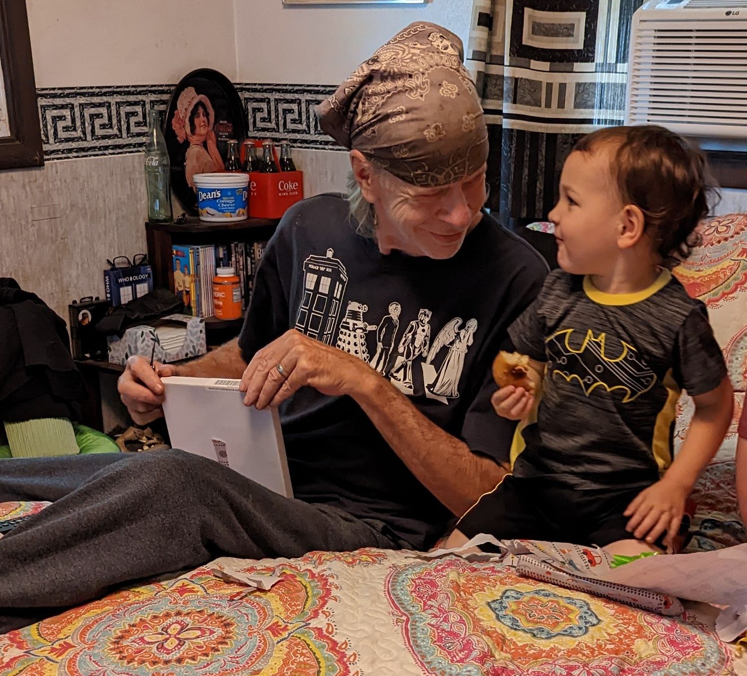 Man reading tablet to curious toddler in Batman shirt while sitting on bed.