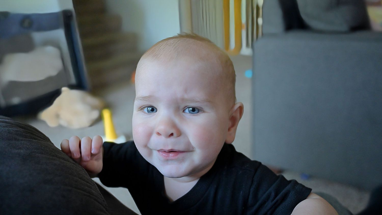 Close-up of smiling baby in black shirt leaning against gray sofa.