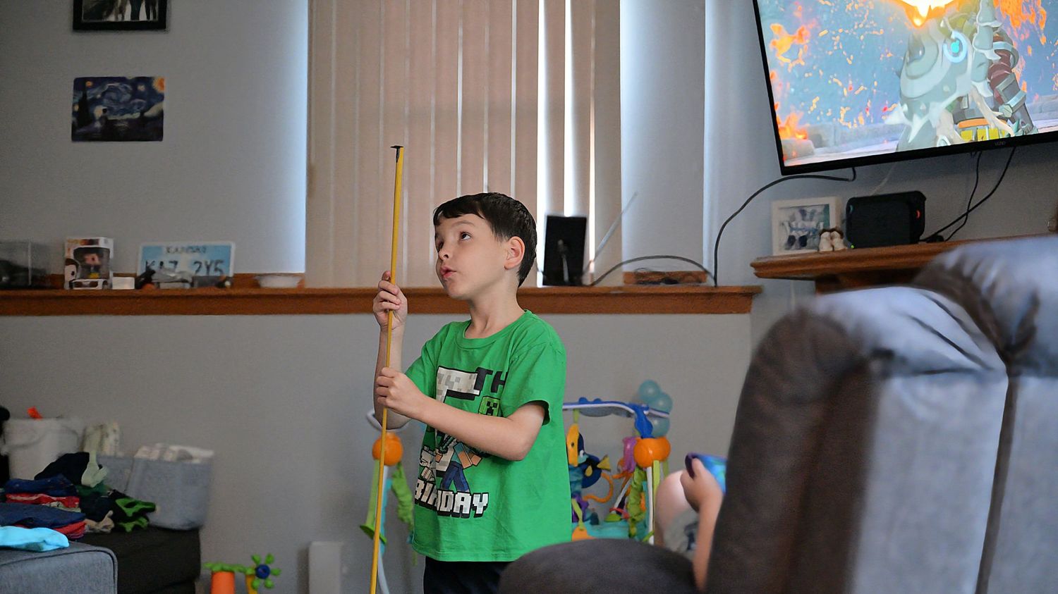 A young boy in a green shirt plays with a yellow toy wand in a cozy living room with a TV in the background.