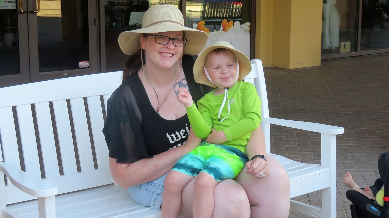 Woman wearing sun hat sits on white bench holding toddler in green outfit outside a shop.