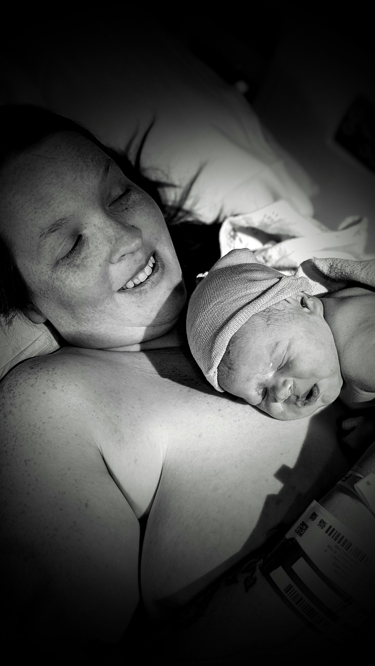 Black and white photo of smiling new mother lying in hospital bed with newborn baby on her chest.