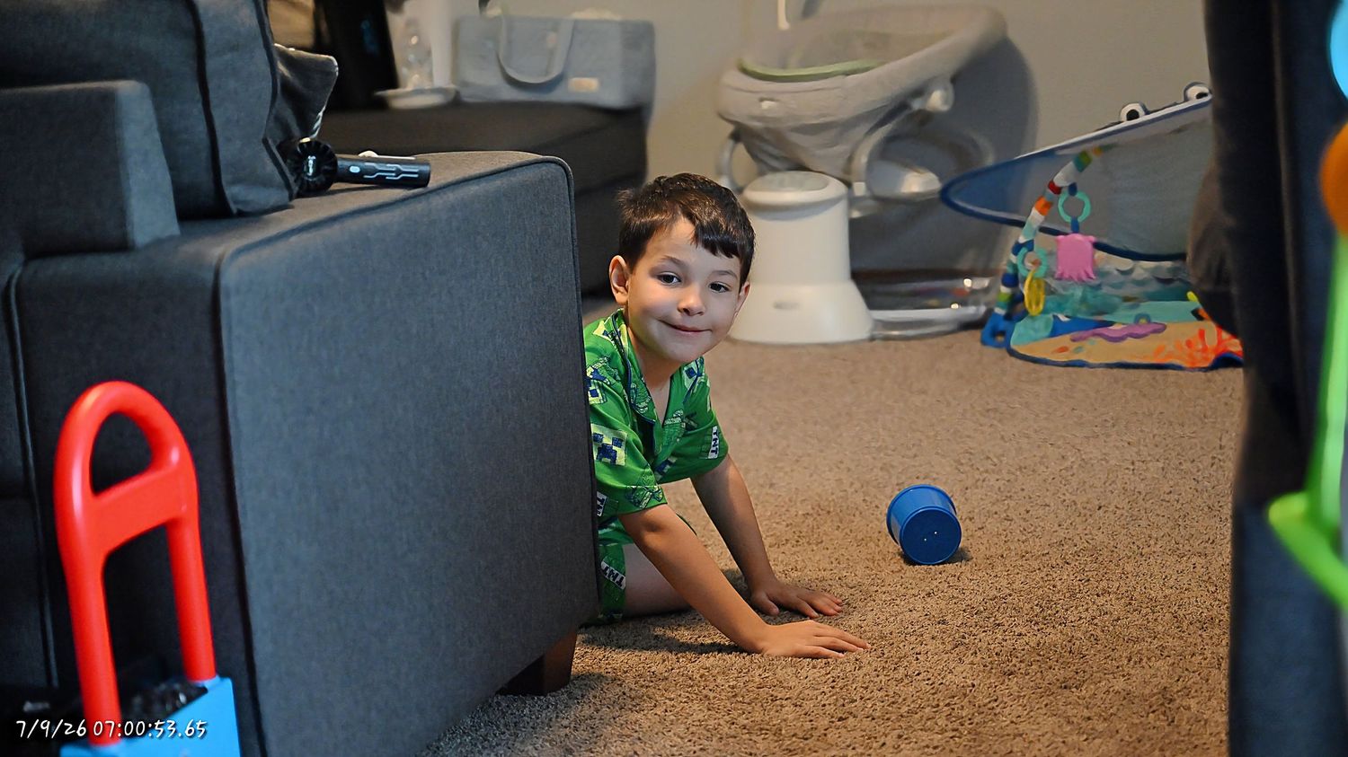 A smiling young boy in green pajamas hiding behind a couch on a carpeted floor with toys scattered around.
