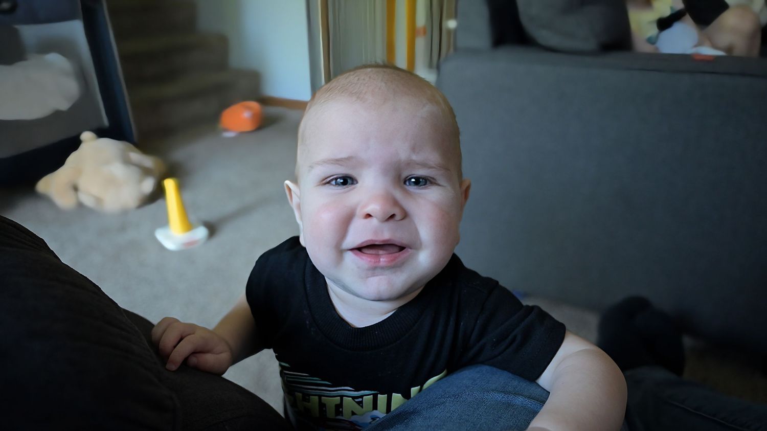 Crying baby in black graphic tee leaning against gray sofa indoors.