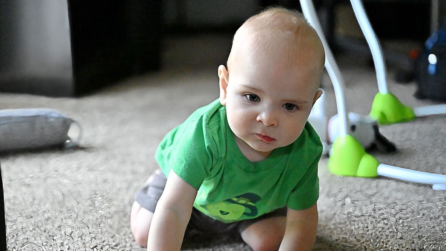 Baby in a green shirt crawling on carpet floor at home.