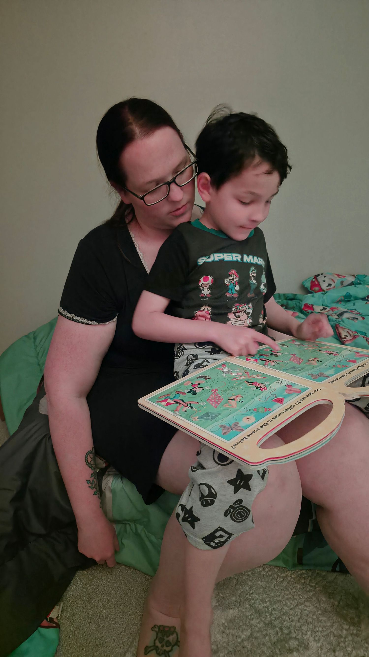 A mother wearing glasses reads a colorful book with her young son on a bed.