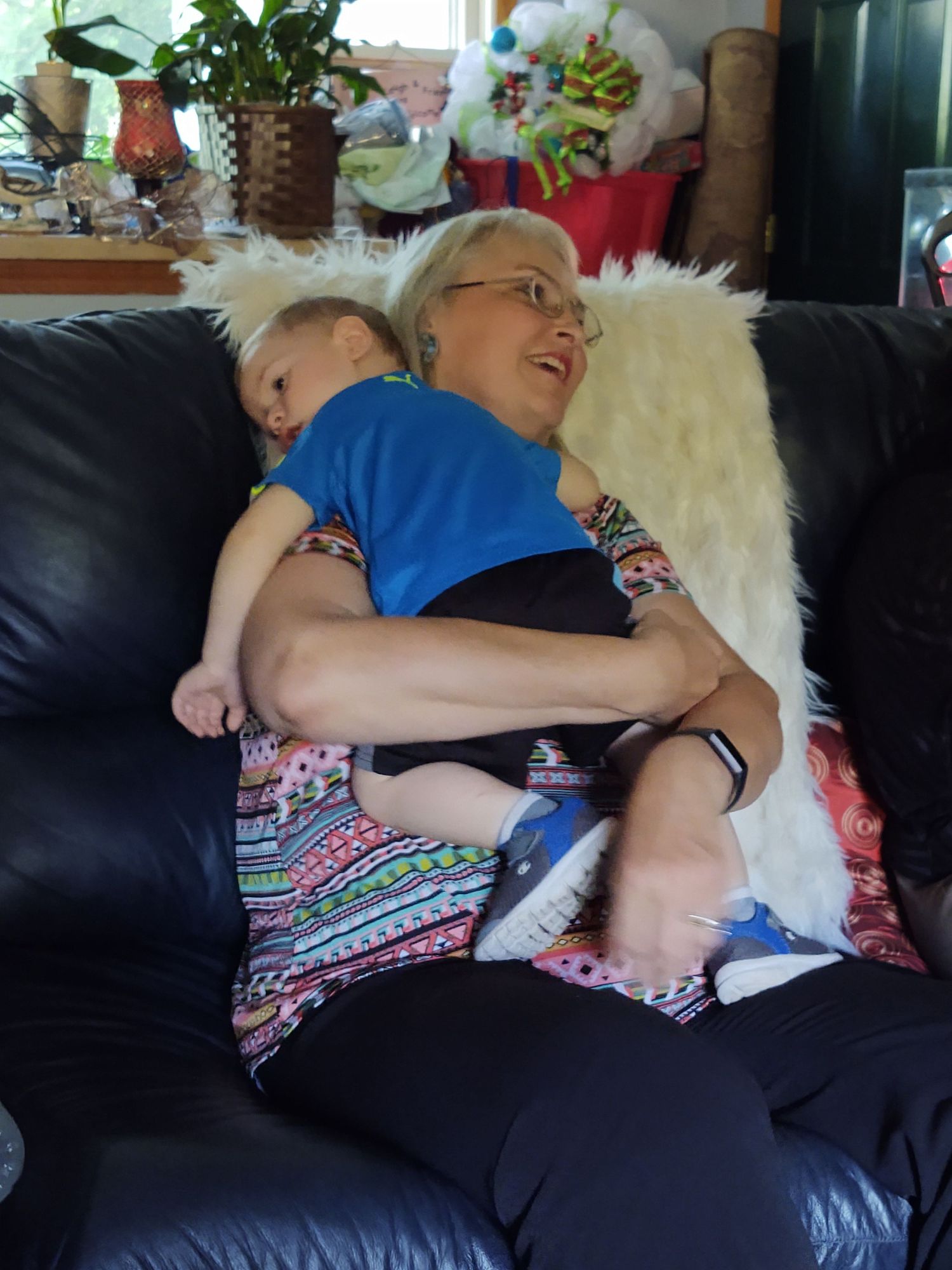 Grandmother sitting on black leather sofa holding sleeping baby in blue shirt.