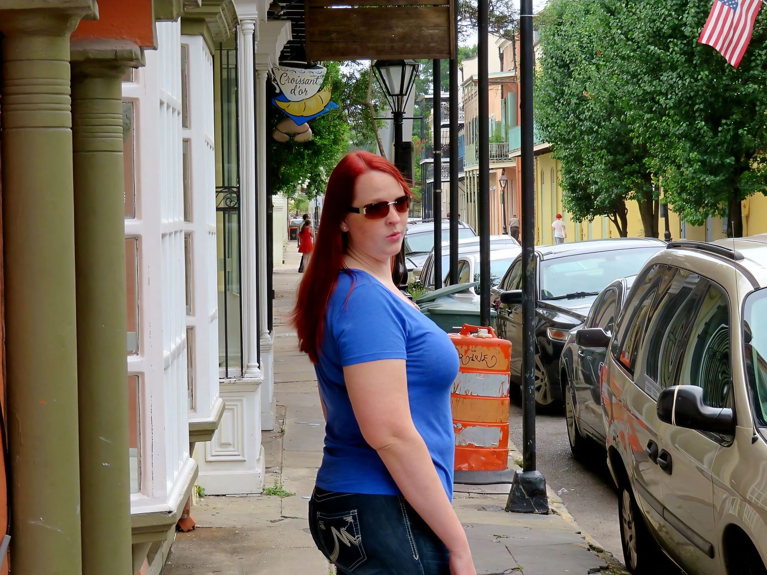 A red-haired woman in a blue shirt and sunglasses standing on a city sidewalk.
