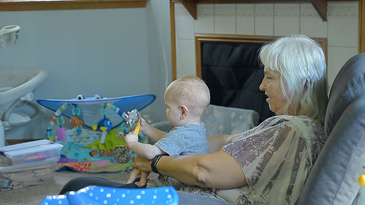 An elderly woman sits with a baby on her lap, engaging with a colorful play mat.