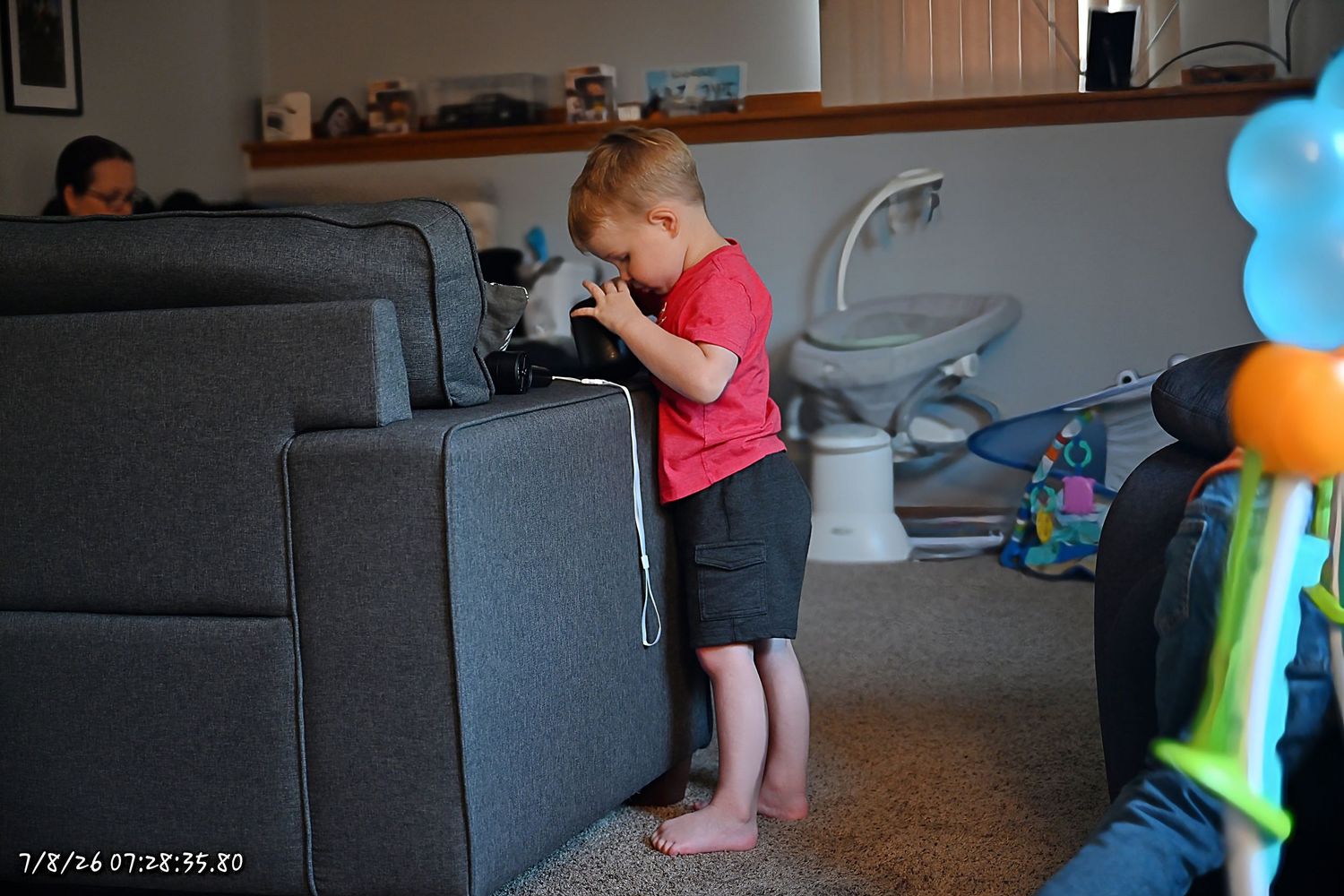 A young boy in a red shirt plays with a device on the side of a gray sofa.