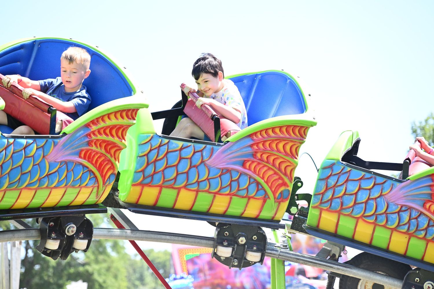 Two children ride a colorful dragon roller coaster at an outdoor carnival.