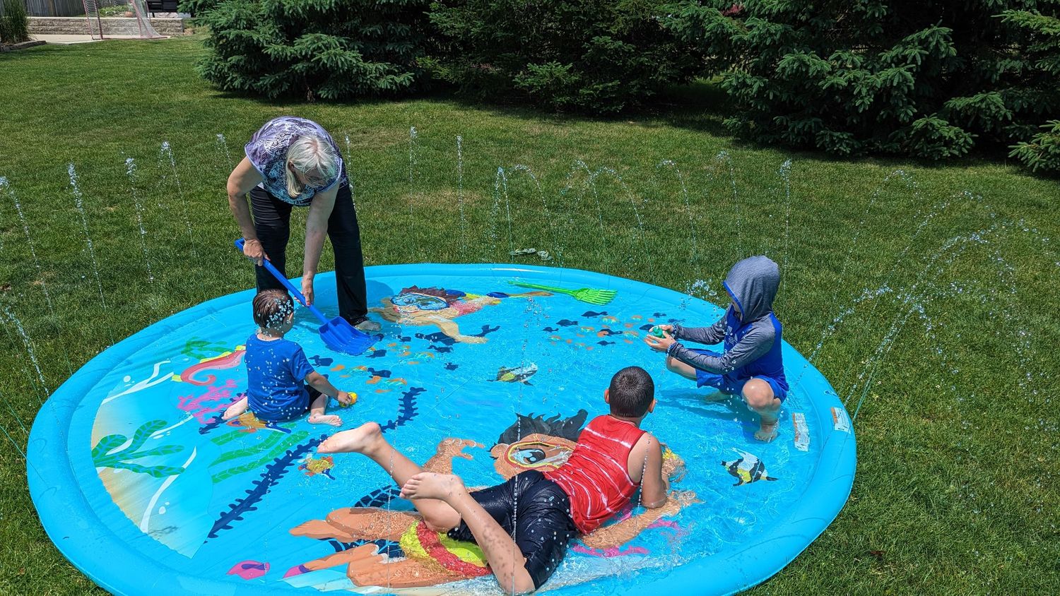 Children playing on blue inflatable splash pad sprinkler mat in sunny green backyard.