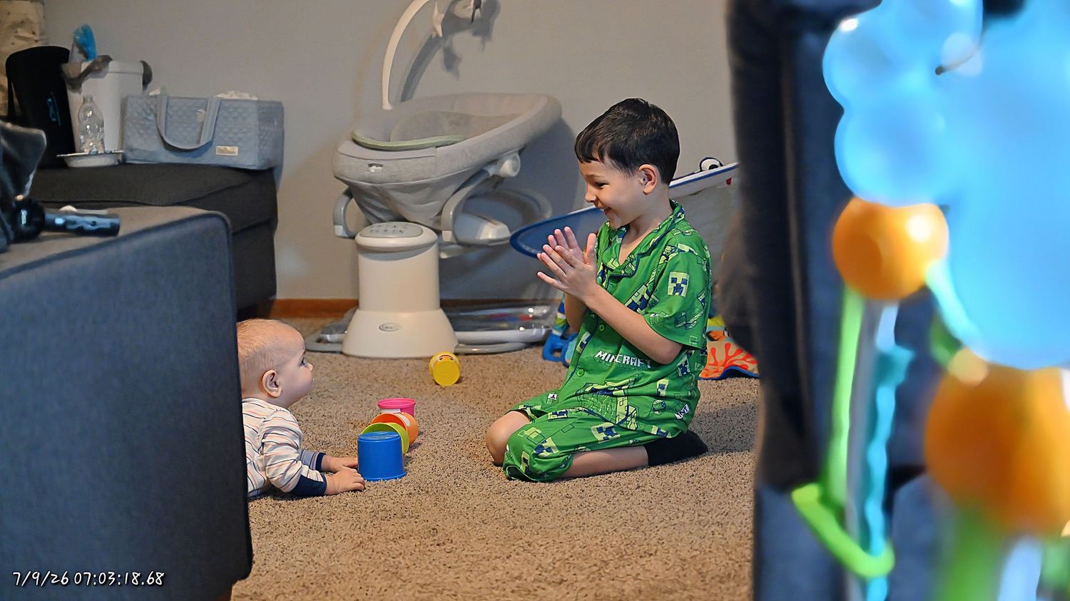 A boy in green pajamas clapping hands while entertaining a baby sitting on a carpeted floor.