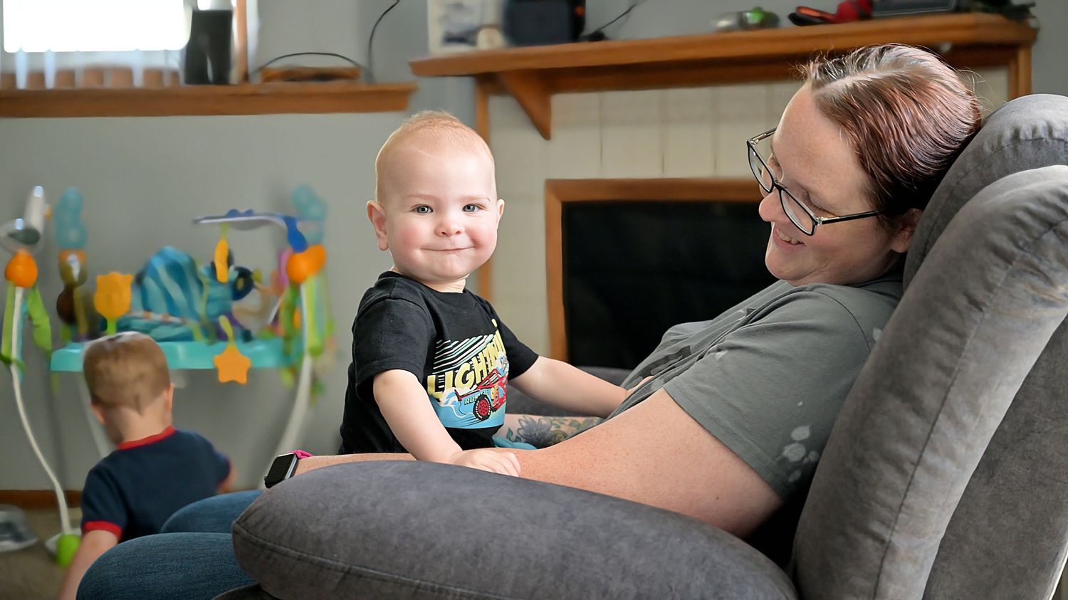 A happy baby sits on a womans lap in a gray recliner while a child plays in the background.