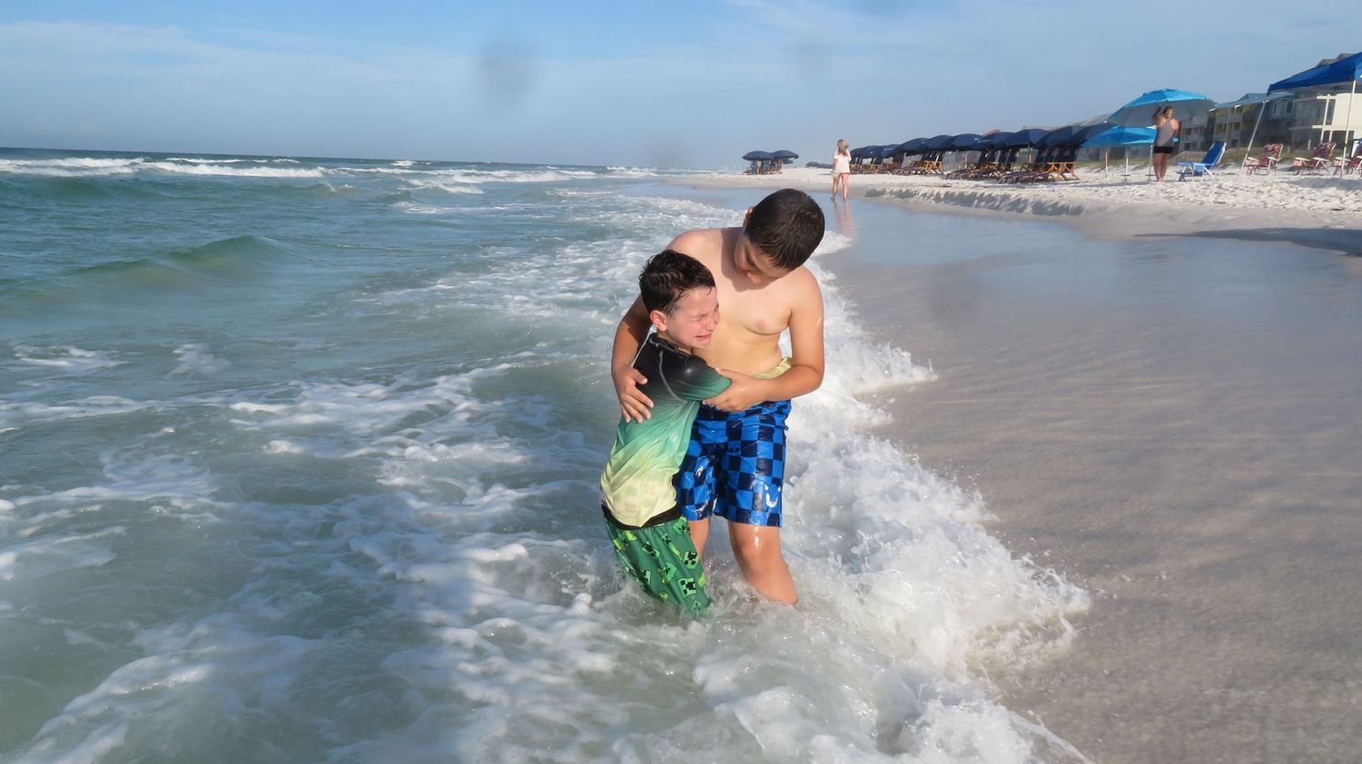 Two young boys embrace and play together in shallow ocean waves on a bright sunny beach day.