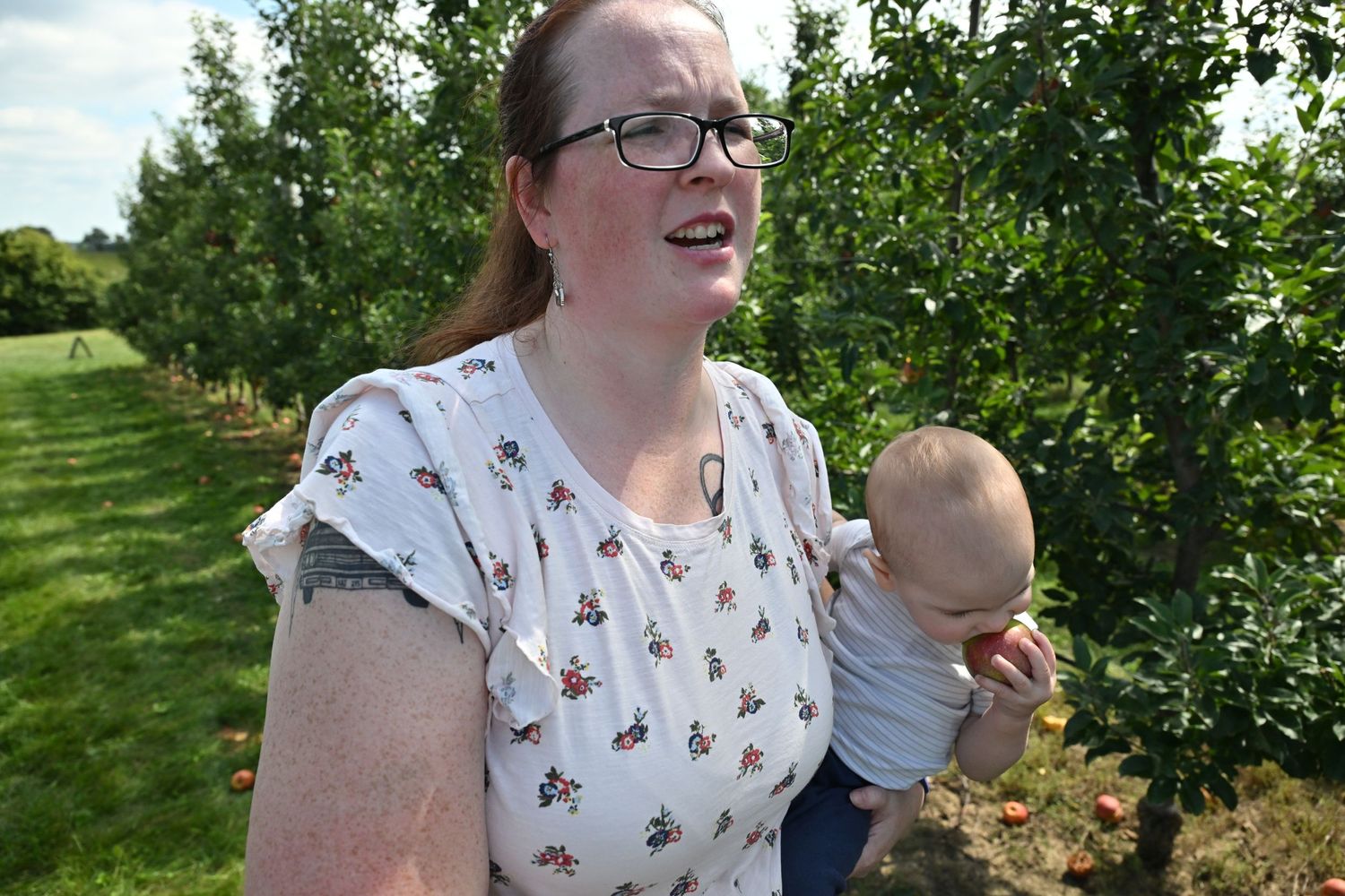 A smiling woman with glasses holds a baby while picking apples in an orchard.