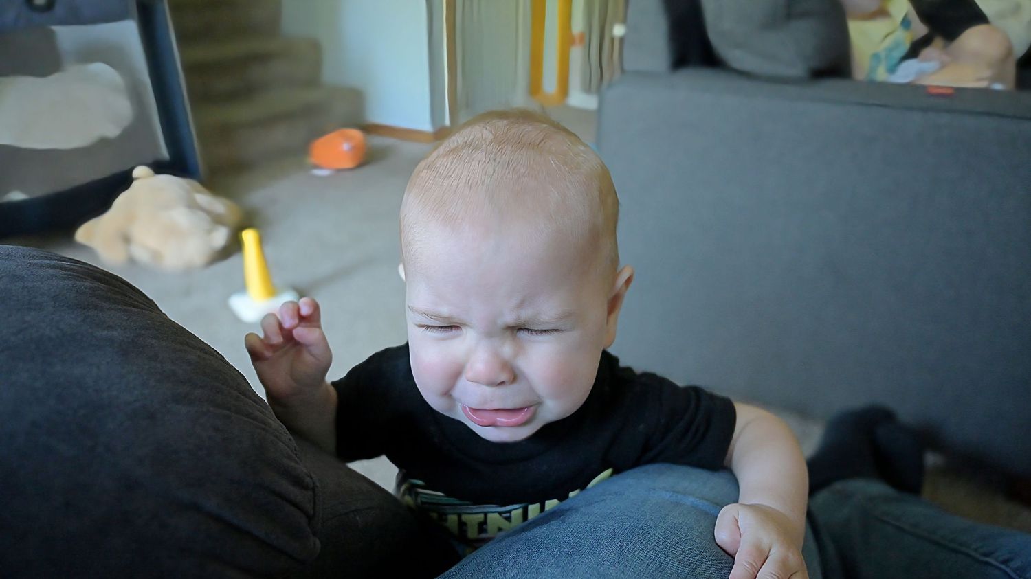 Upset baby in black shirt crying while being held near gray sofa.