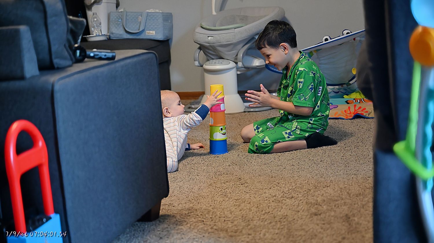 A young boy in green pajamas stacking colorful cups with a baby lying on a carpeted floor.