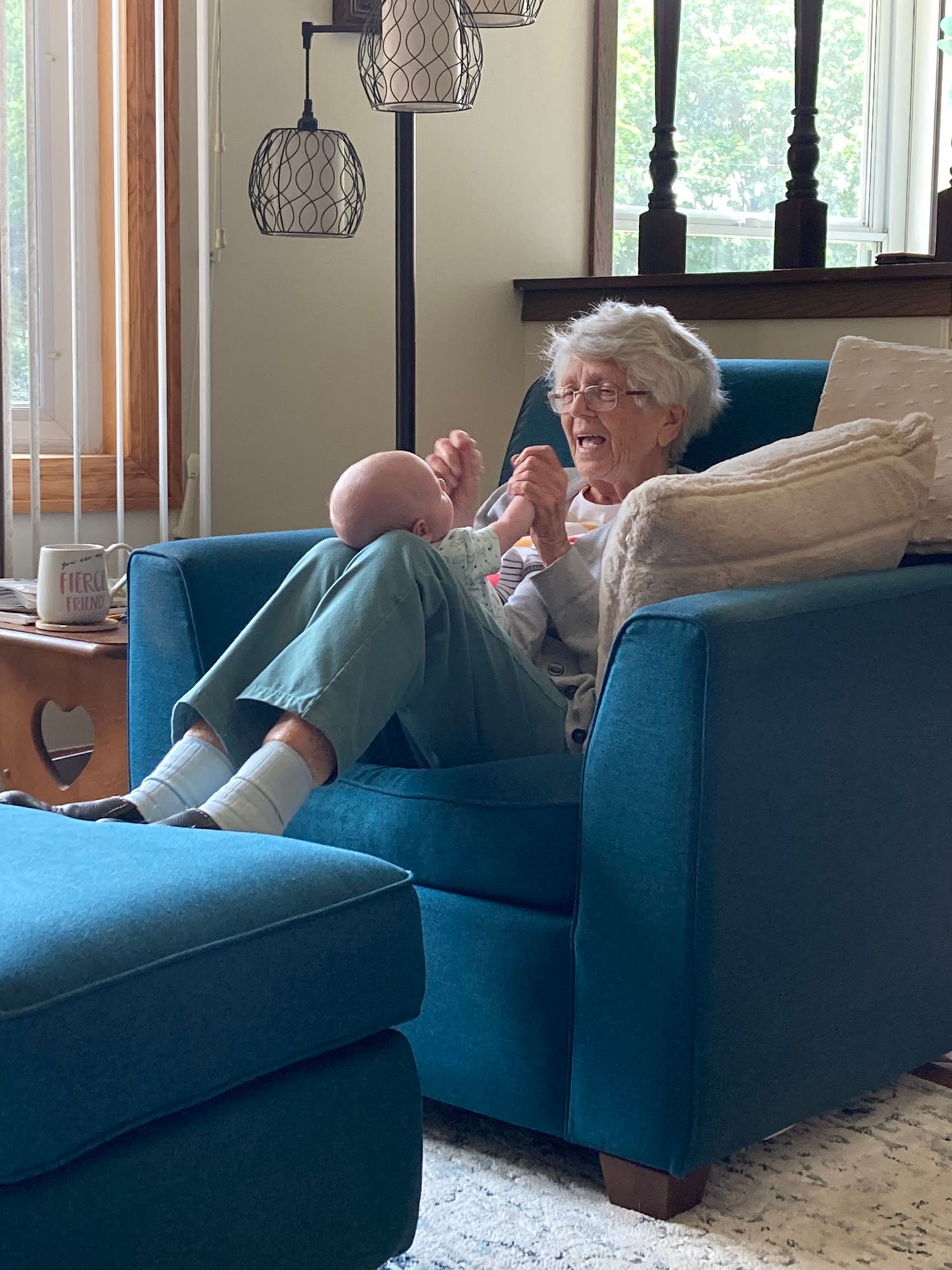 Elderly woman sits in teal armchair smiling while holding a newborn baby in bright living room.