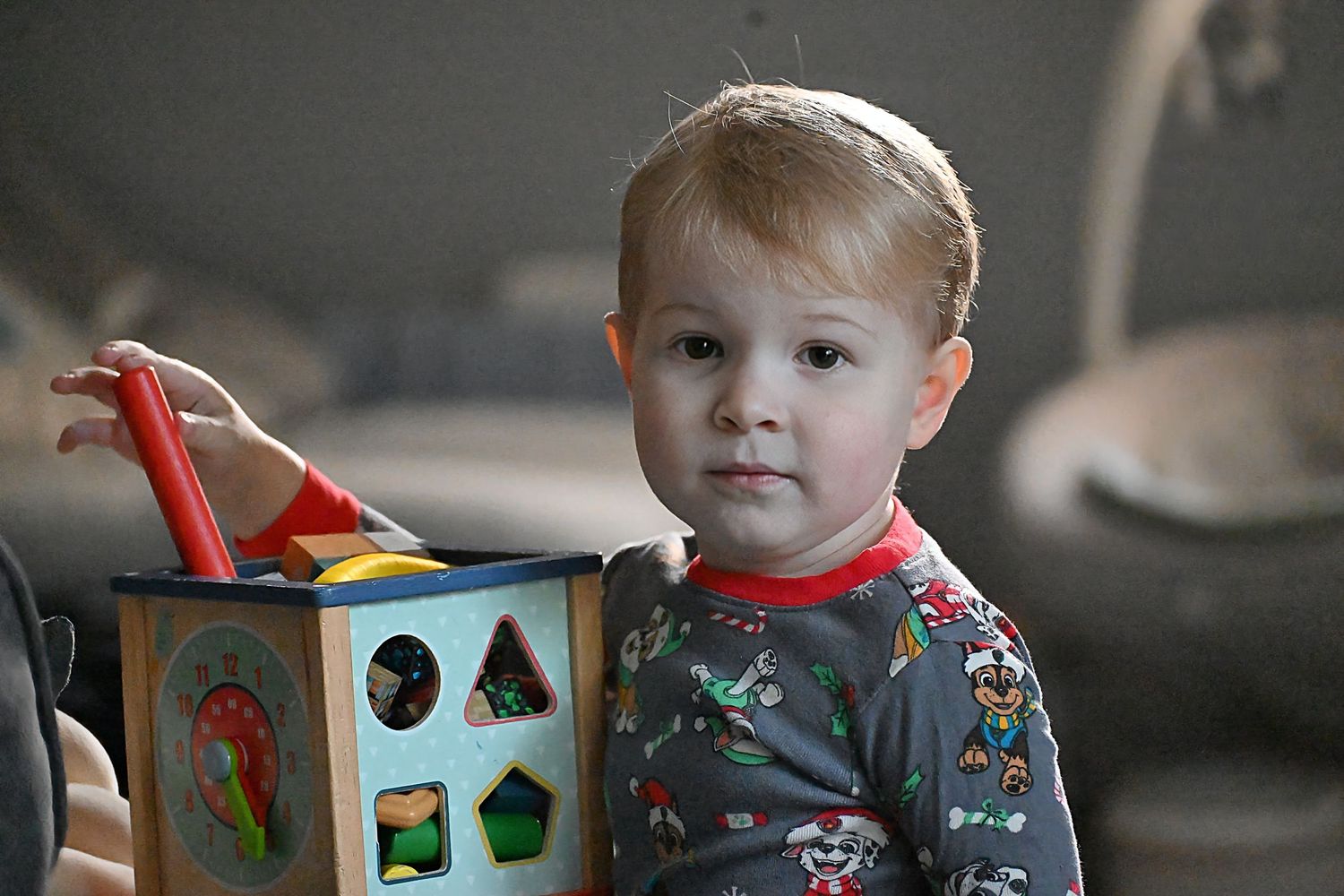 A toddler in pajamas plays with a wooden shape-sorting cube toy indoors.