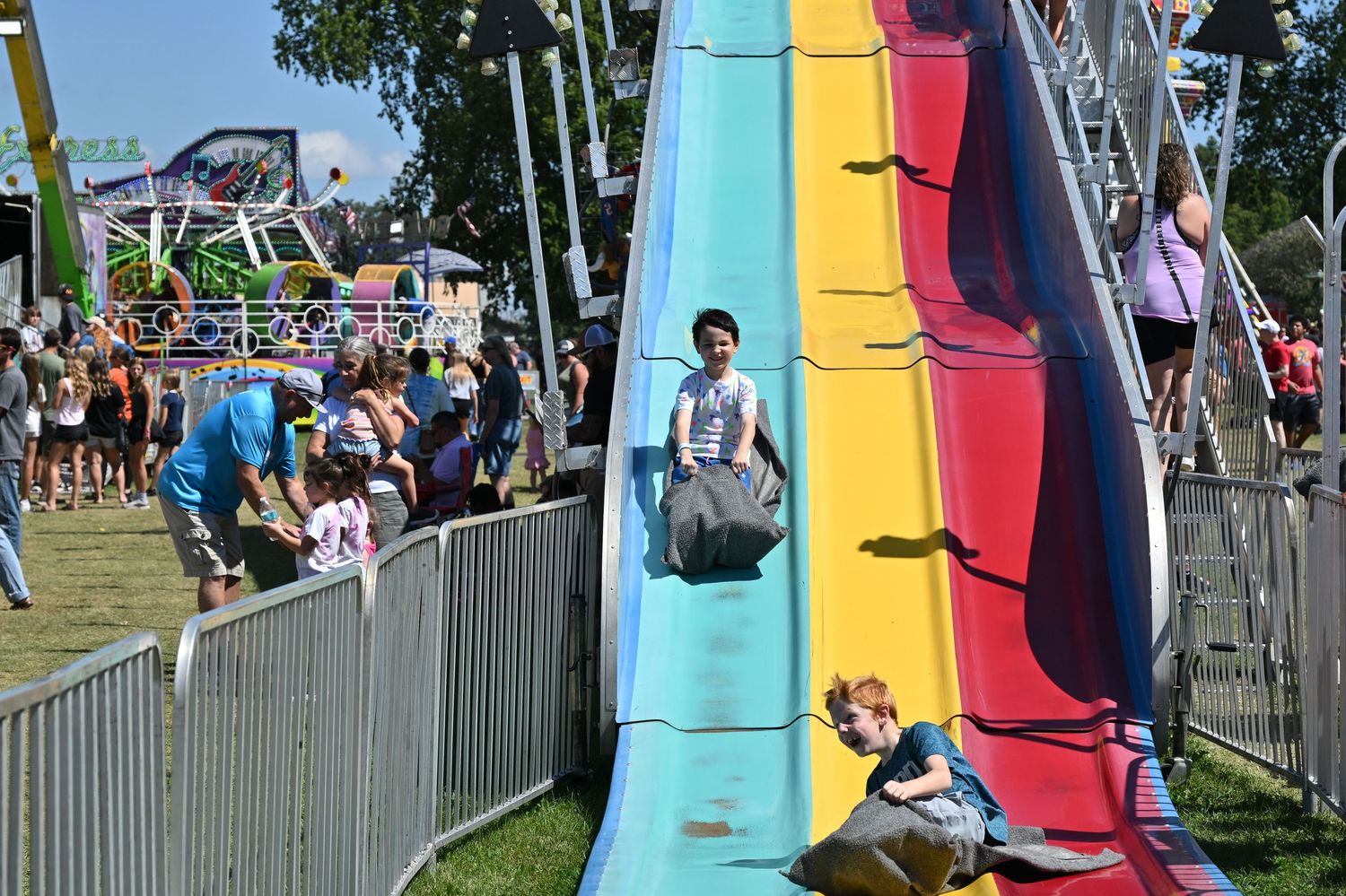 Children slide down a colorful giant slide at a sunny outdoor carnival.