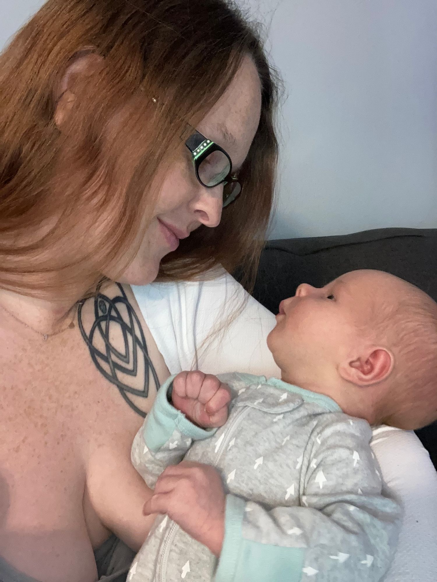 Red-haired tattooed mother with glasses tenderly gazes at her newborn baby in her arms.