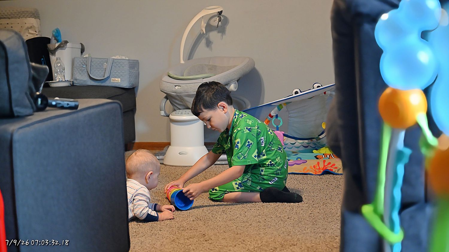 An older boy in green pajamas playing with a baby on the floor surrounded by colorful toys.
