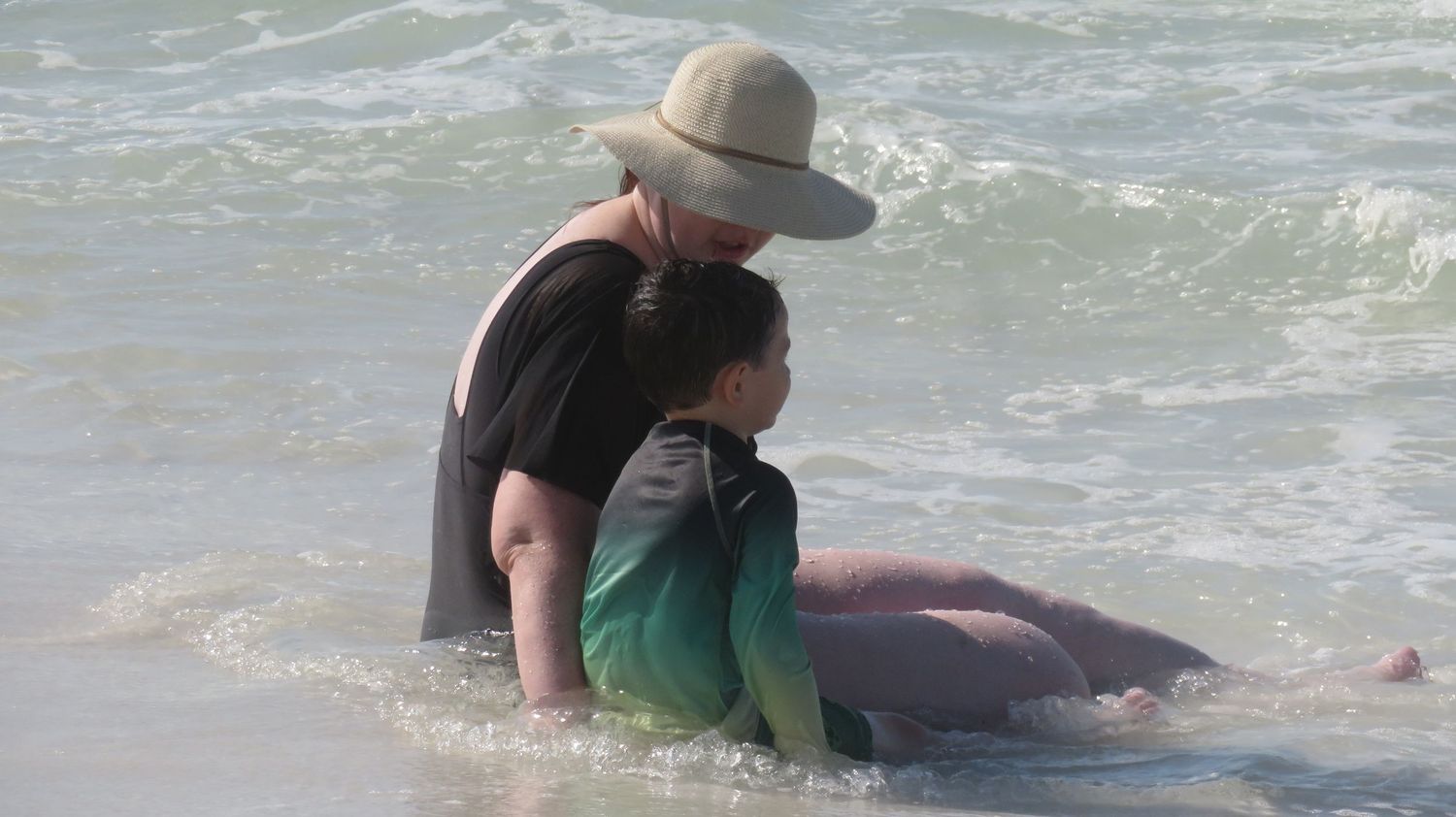 Woman in black swimsuit and sun hat embraces child in green rash guard in ocean waves.
