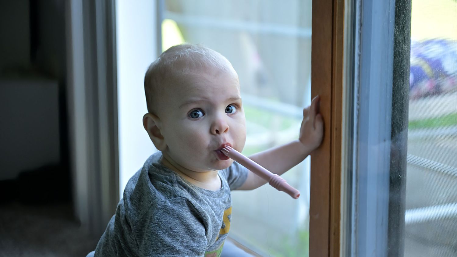 A baby stands at a glass sliding door, chewing on a stick while looking outside curiously.