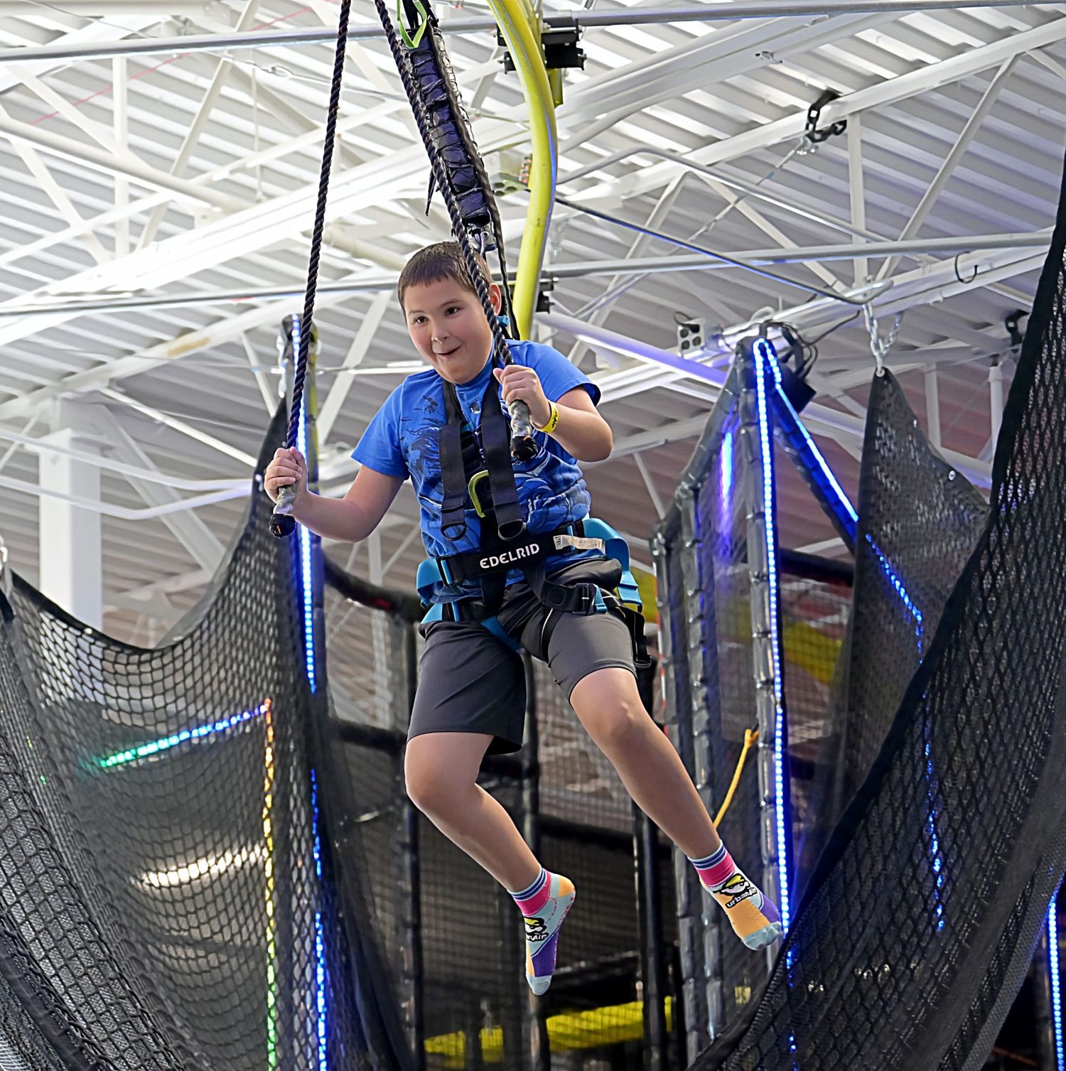 A smiling child in a blue shirt enjoys a zip line ride at an indoor adventure park.