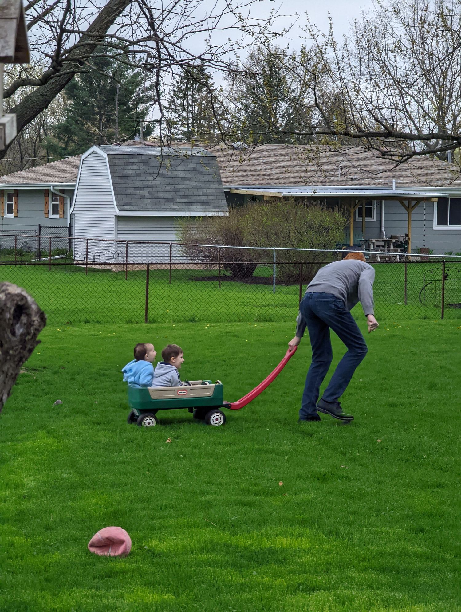 Father pulling two young children in green wagon across lush backyard lawn.