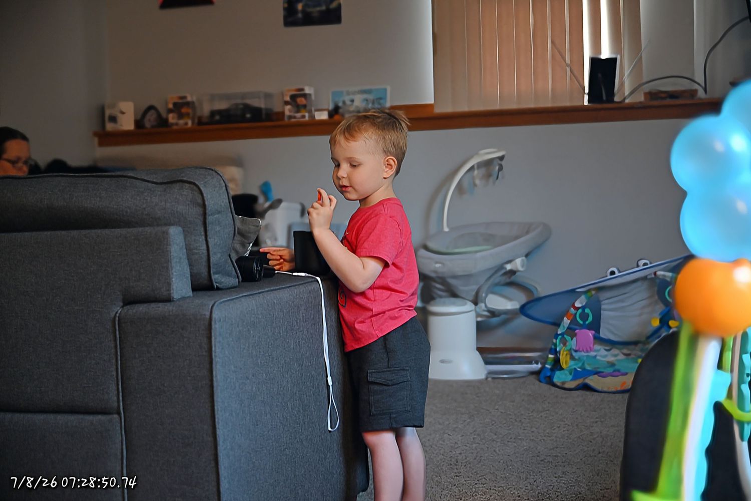 A young boy in a red shirt stands in a living room eating a snack near a sofa.