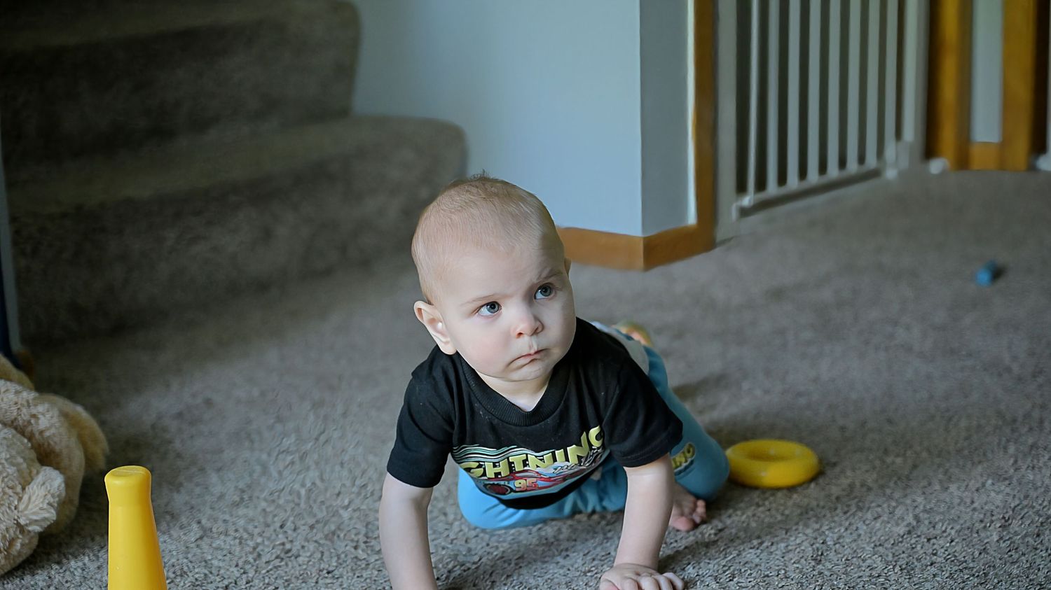 Baby in black graphic tee sitting on carpet near staircase with yellow toy.
