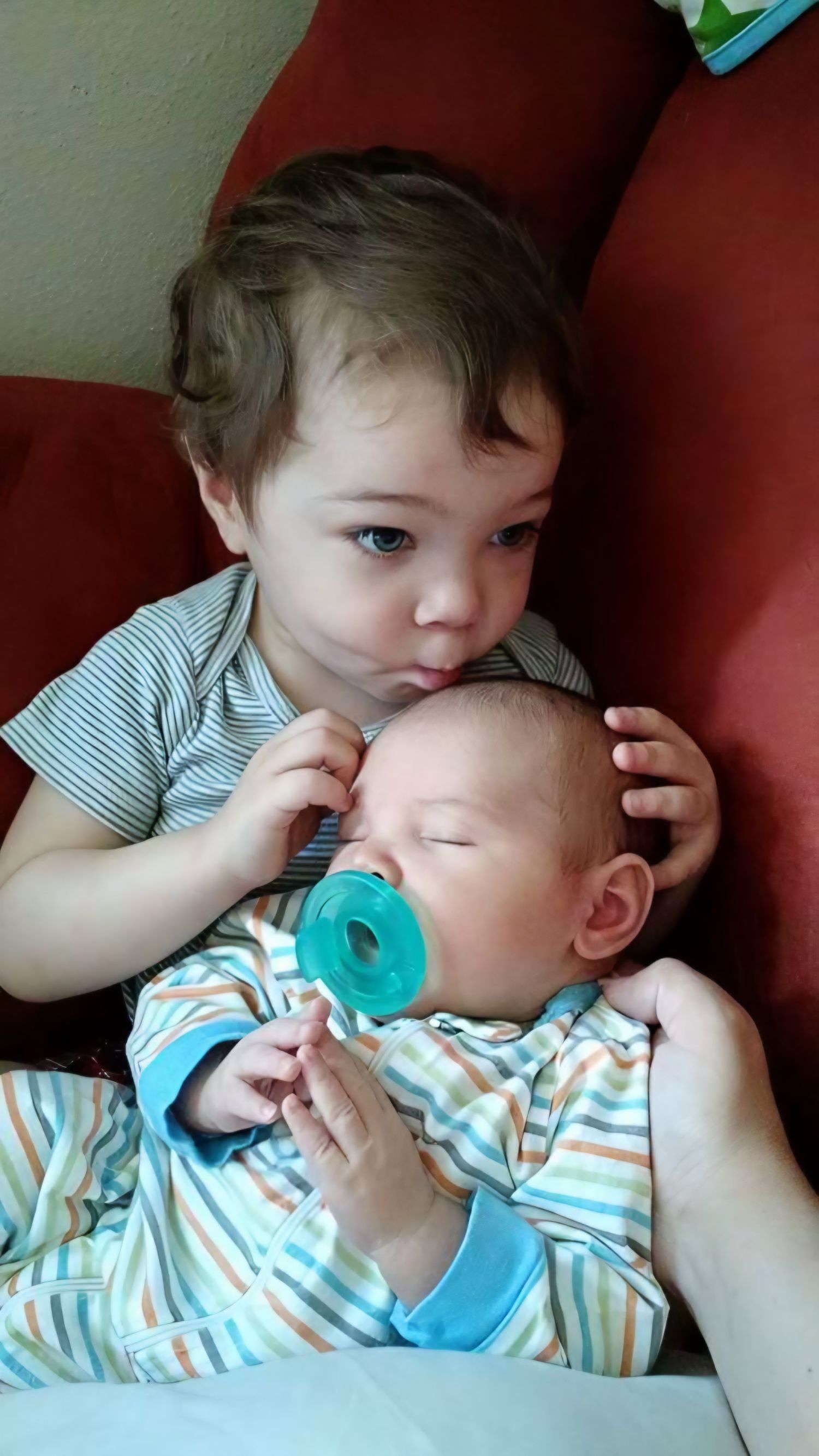 Toddler gently holding newborn sibling with pacifier on a red couch.