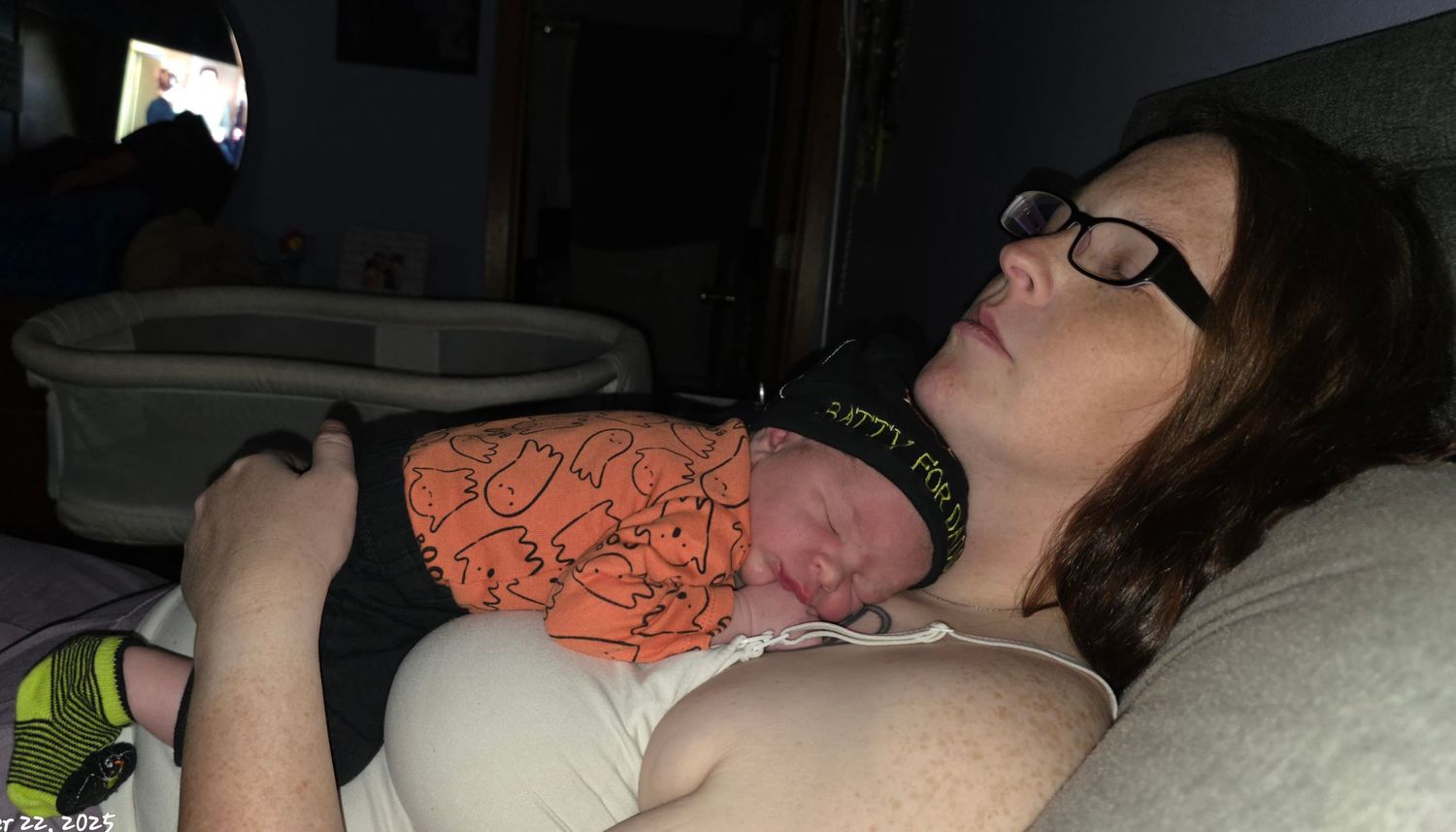A tired mother rests while cradling her newborn baby dressed in an orange outfit.