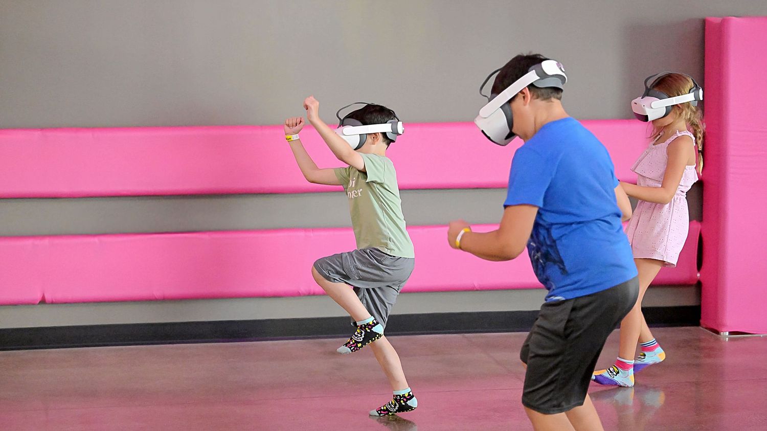 Kids and adult wearing VR headsets playing virtual reality games in a pink-walled room.