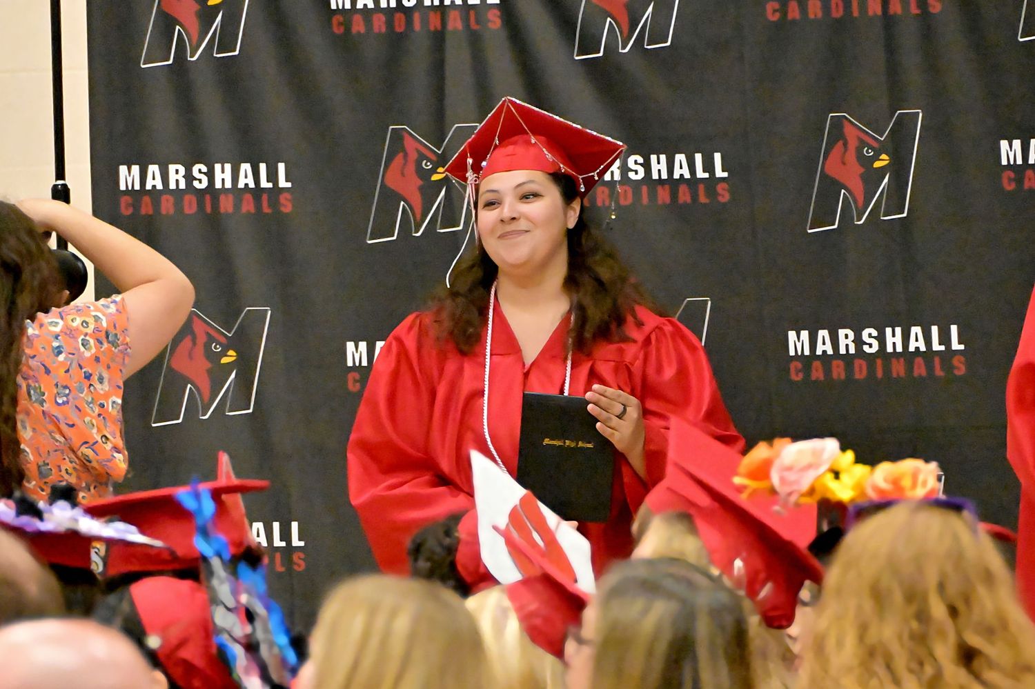 Female graduate in red cap and gown holding diploma at Marshall Cardinals ceremony.