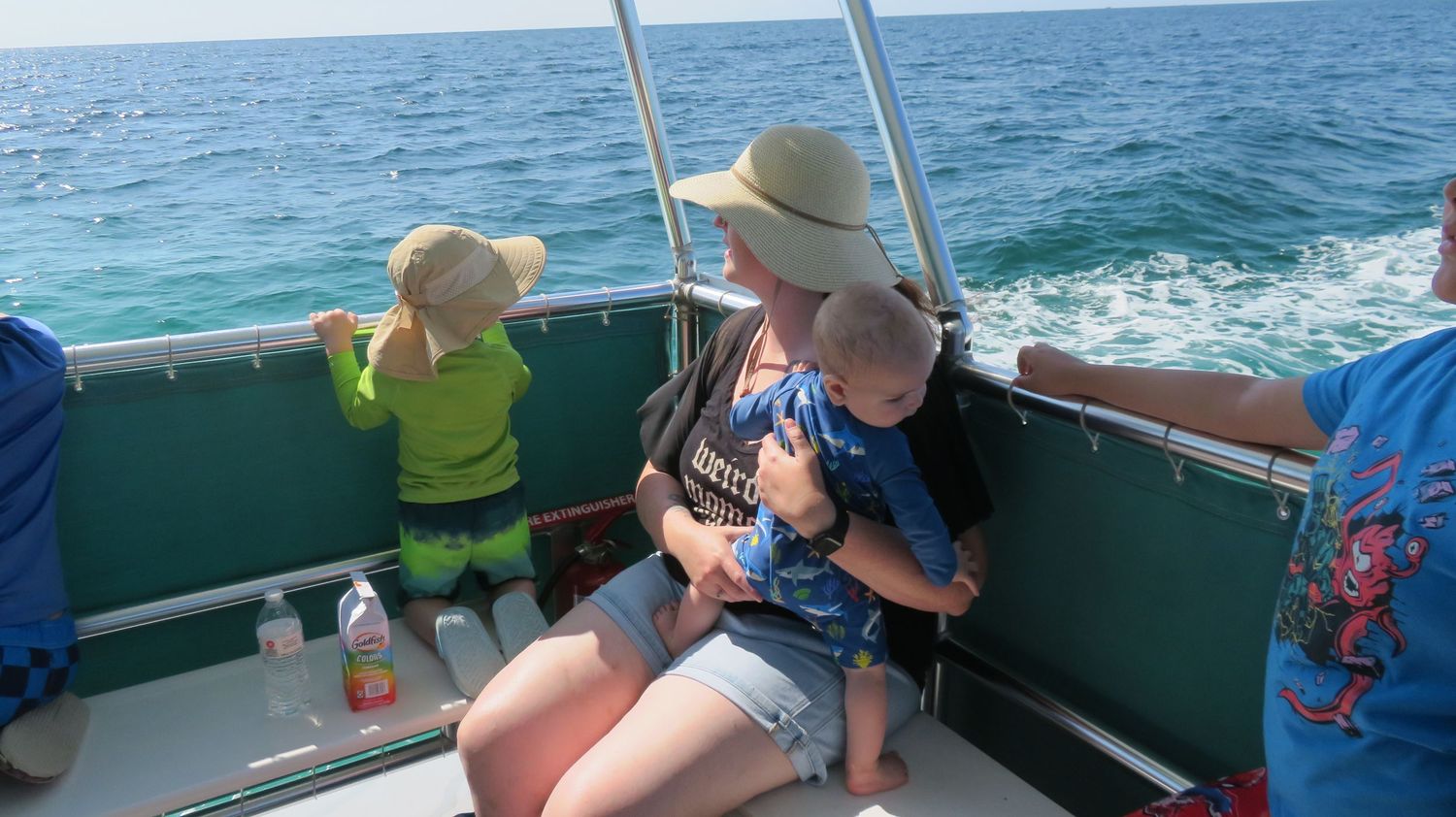 Mother in straw hat holding infant on a boat while toddler looks out at the ocean.