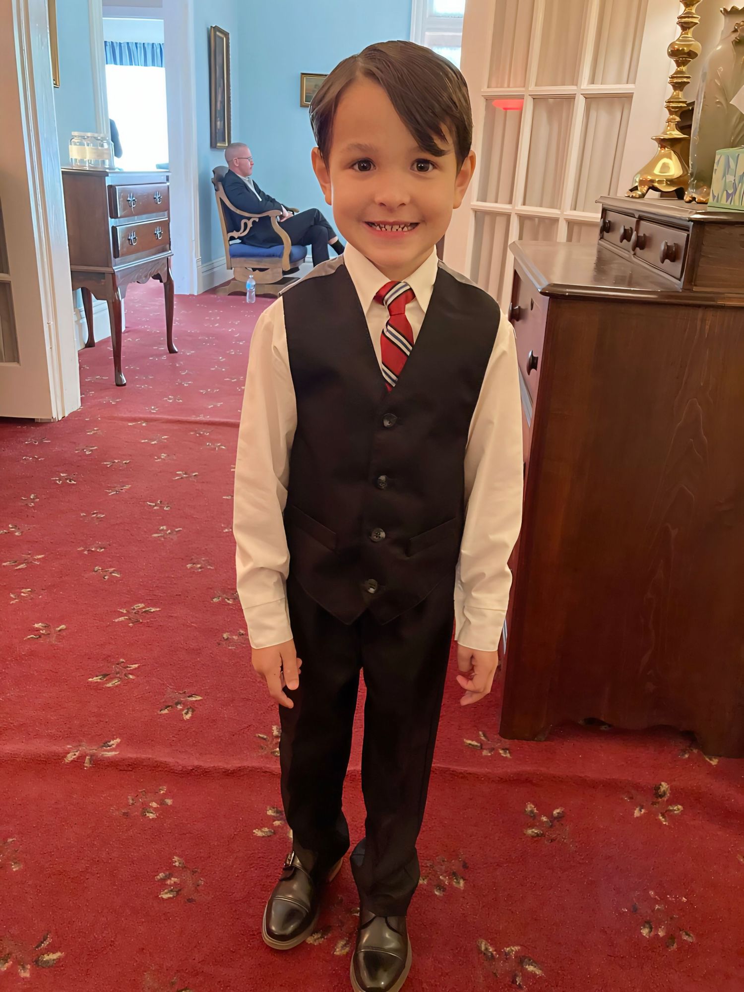 A young boy smiles in a formal black vest, white shirt, and red tie on a red carpet.