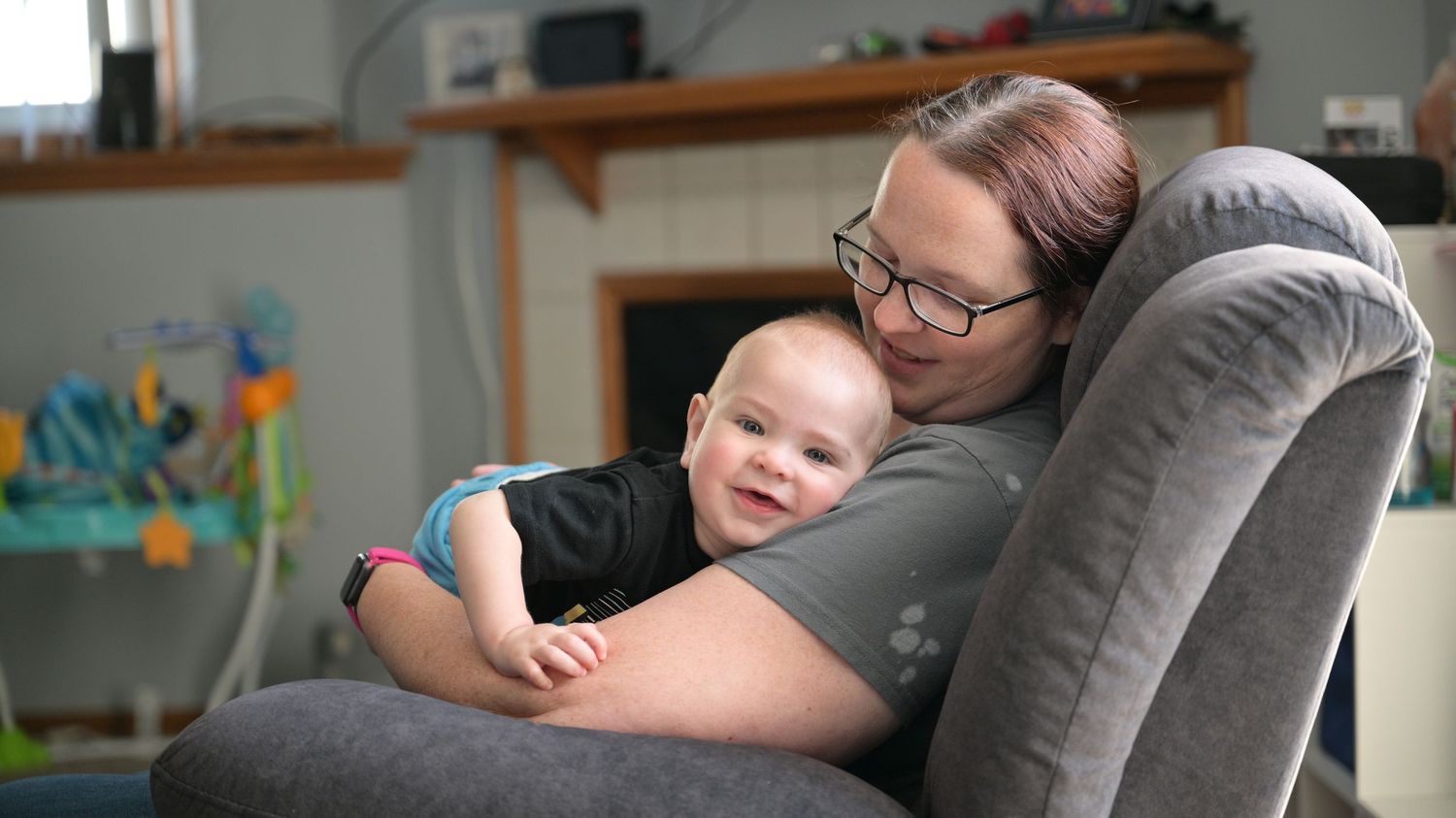 A mother with glasses smiles warmly while cuddling her happy baby in a gray armchair.