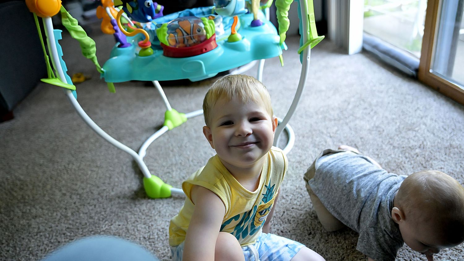 A smiling toddler reaches toward the camera beside a colorful baby activity jumper toy.